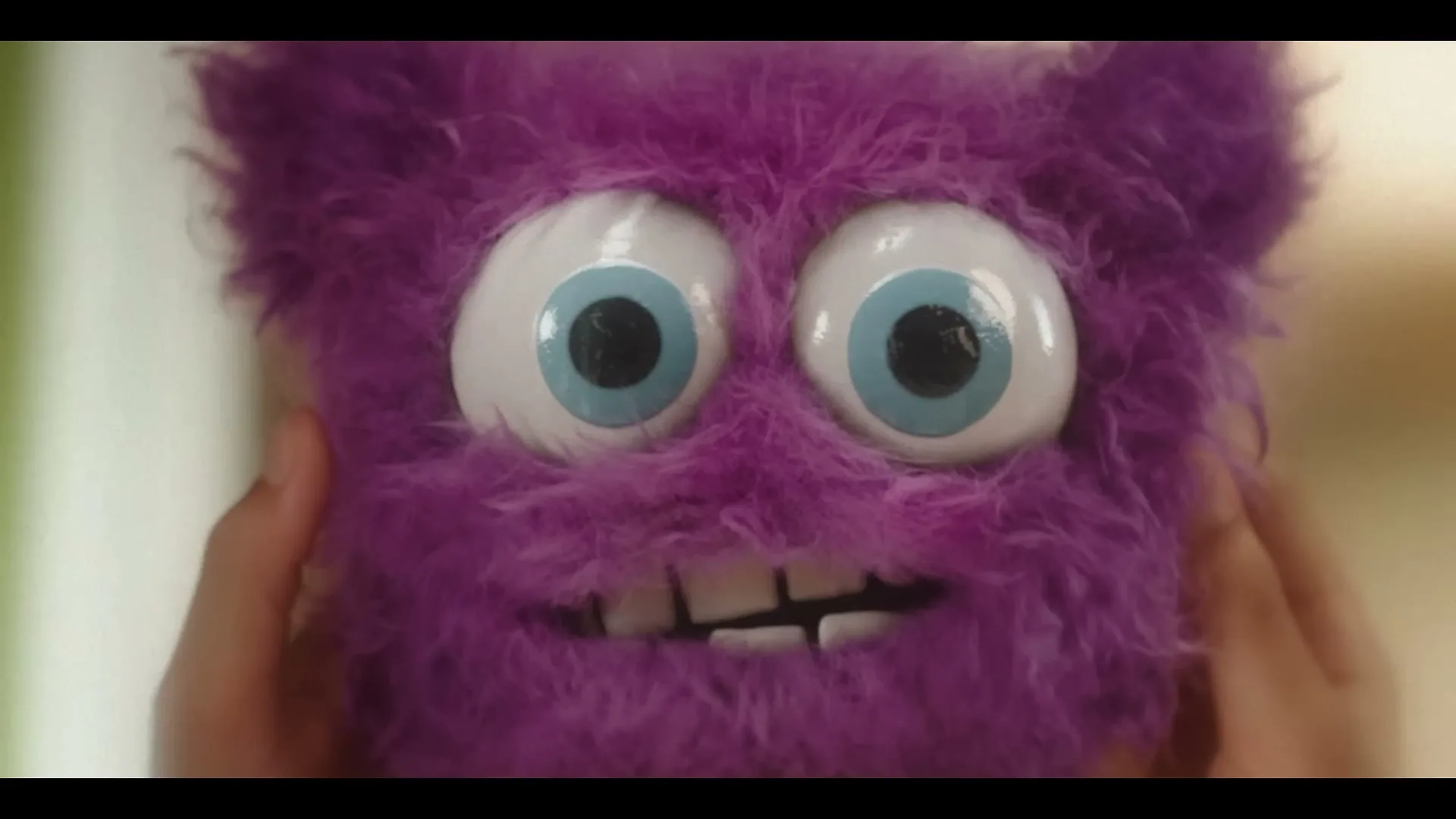 A child's hands hold a purple, furry monster puppet with large blue eyes and a wide smile, looking directly at the camera.