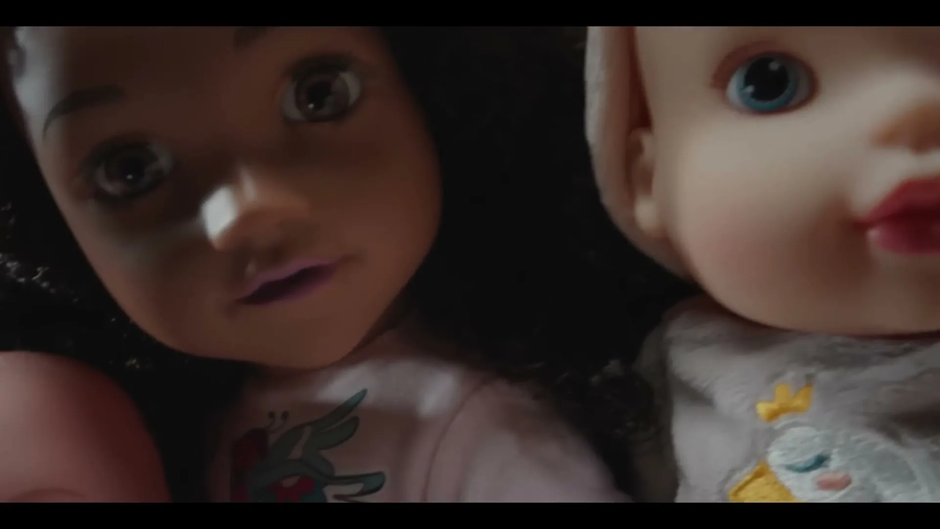 A closeup shot of three dolls, one with light skin and blue eyes, another with dark skin and brown eyes, and a third with light skin and blue eyes, are seen in a dimly lit room.