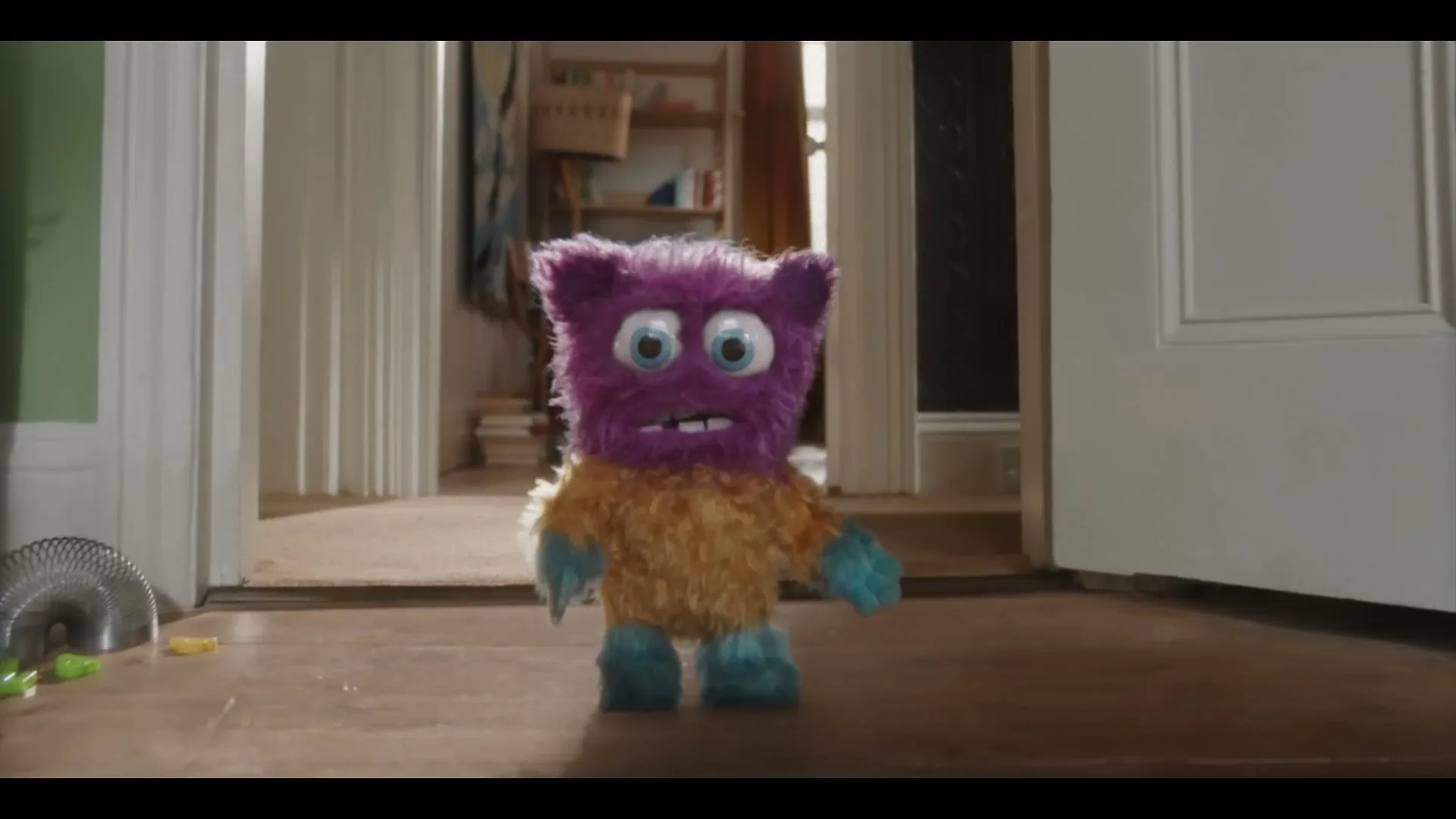 A small, purple and orange furry monster toy stands in a doorway, looking surprised, as a toy car speeds past it on the floor.