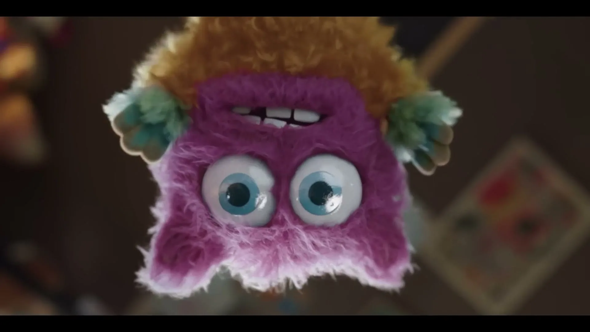A fluffy purple and orange toy monster with big blue eyes hangs upside down, looking directly at the camera in a room.