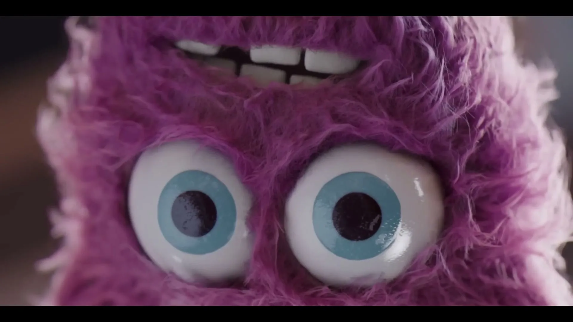 A purple furry monster with large blue eyes and buck teeth looks around, with a green hand on its head, in a close-up shot.