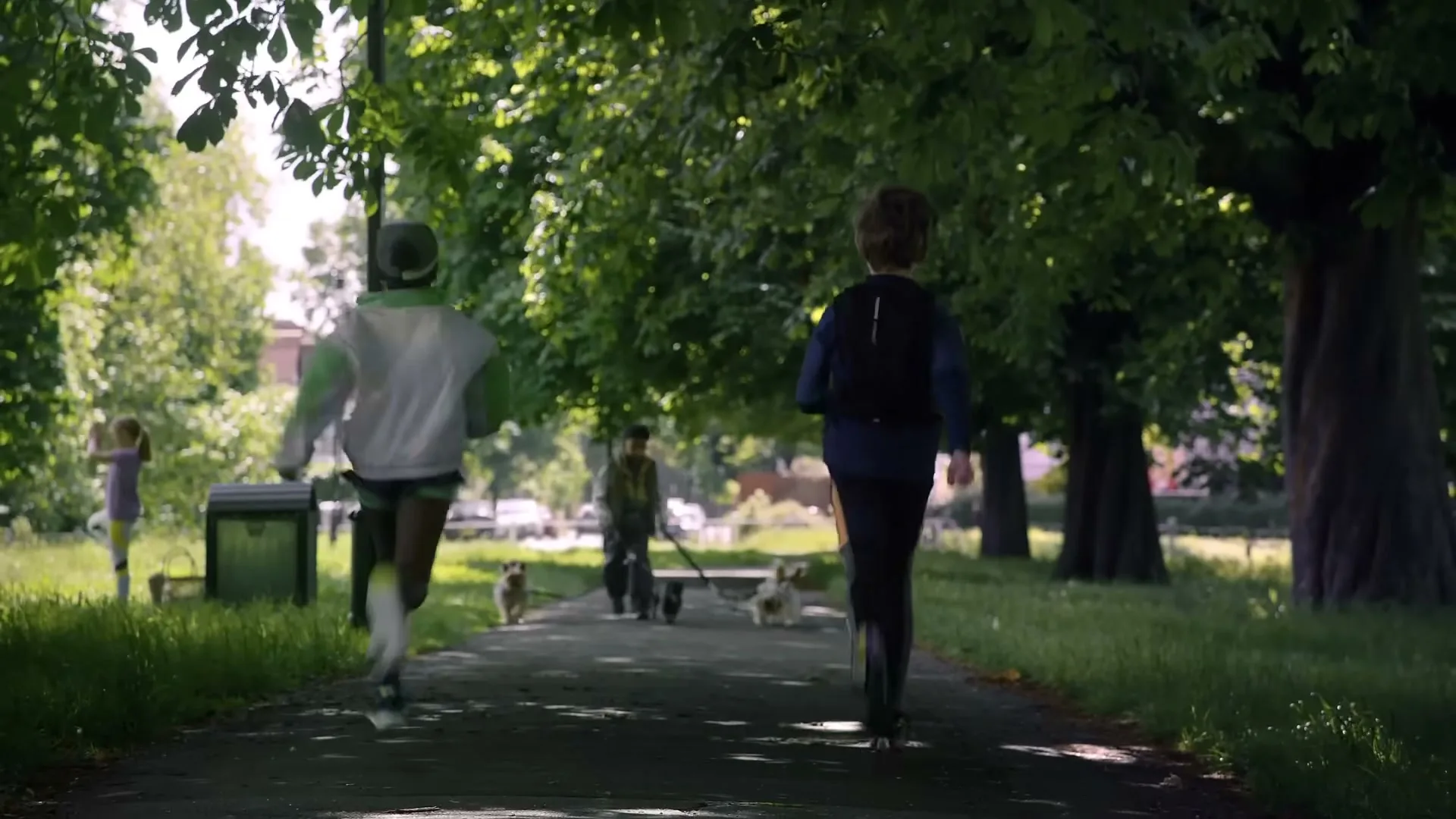 Two children run away from the camera on a path in a park, with other people and dogs in the background.