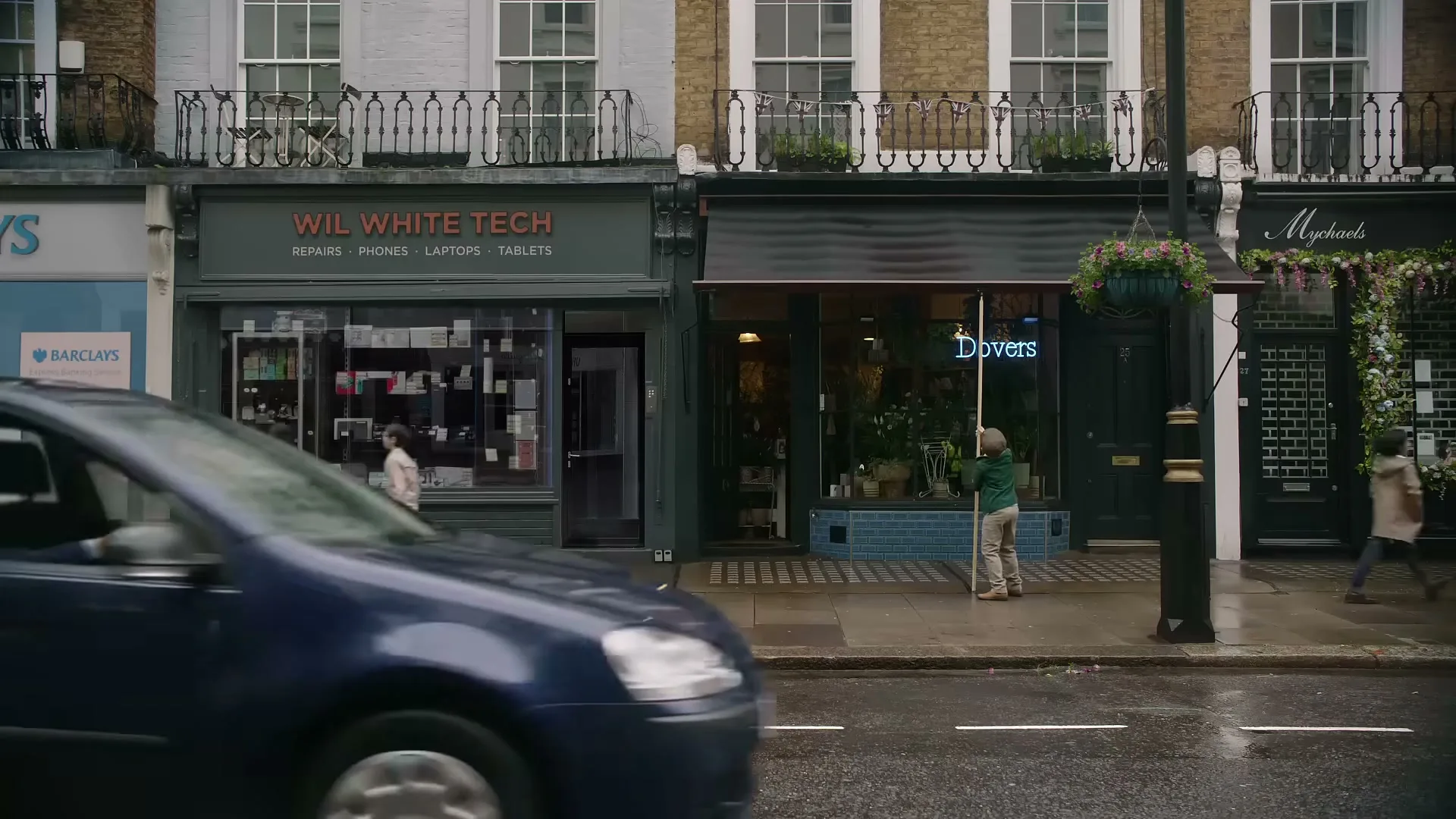 A blue car drives past a row of shops on a wet street, with pedestrians walking on the sidewalk.