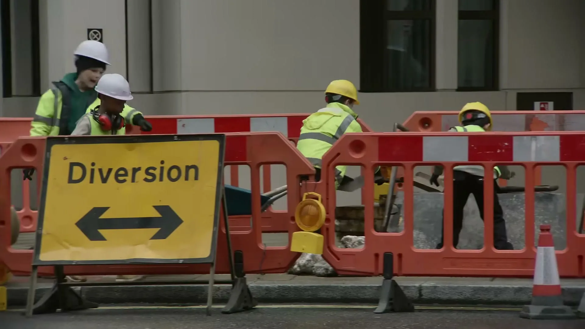 Four children dressed as construction workers are seen behind orange barriers with a yellow "Diversion" sign in the foreground on a city street.