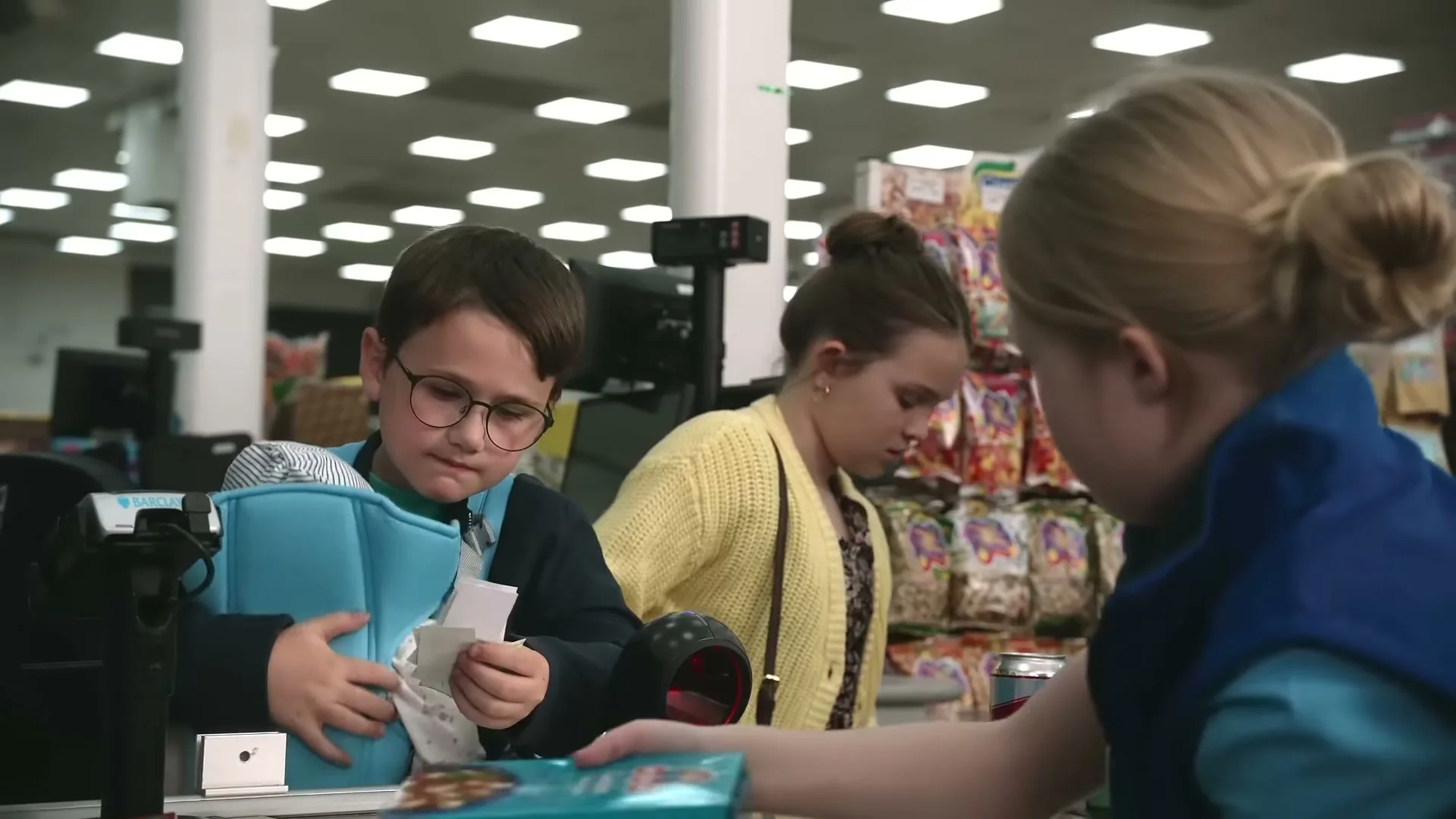 A young boy in glasses, wearing a baby carrier, looks at a receipt while a girl scans items at a grocery store checkout.