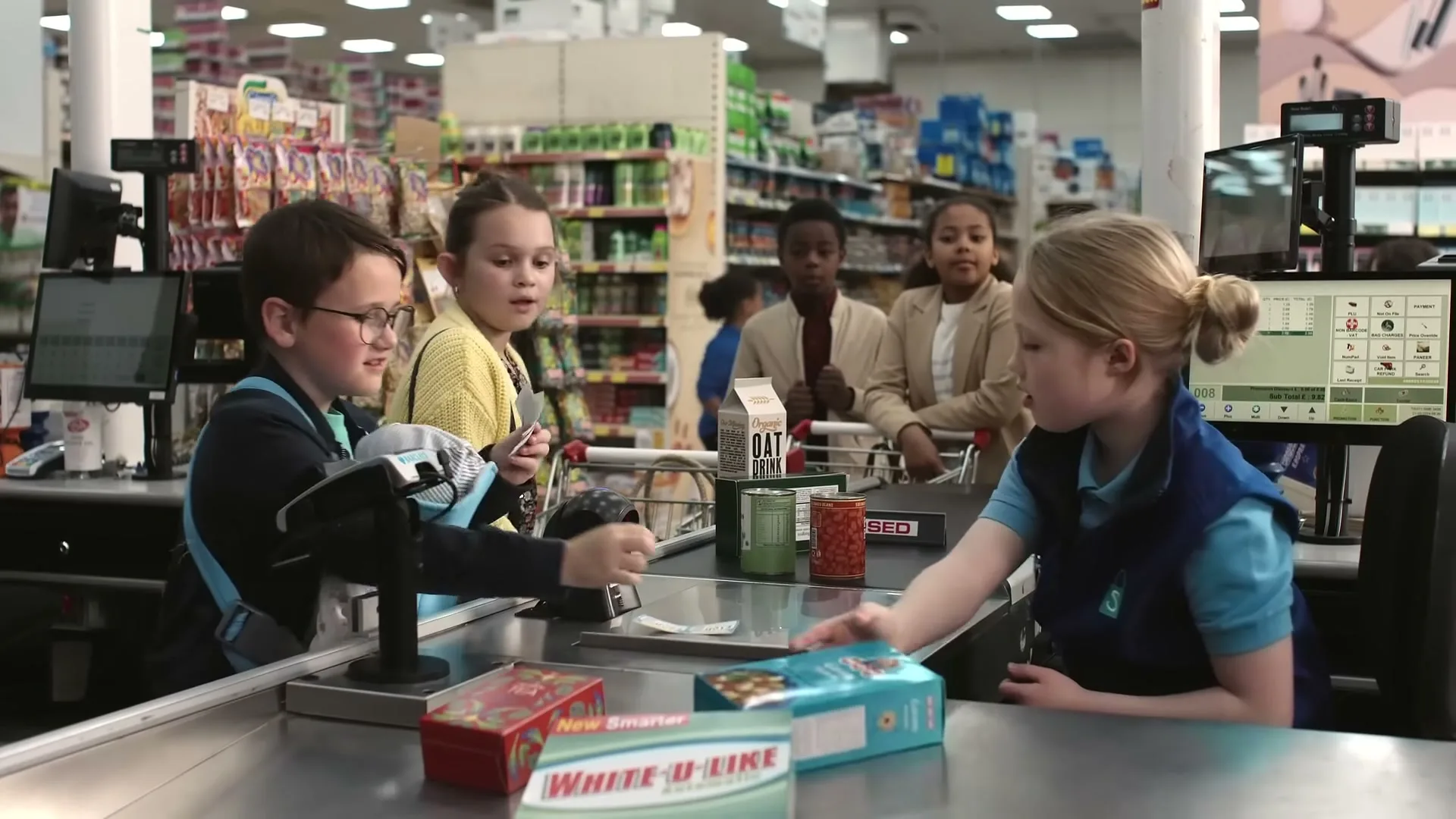 A young boy with glasses and a baby carrier pays for groceries at a supermarket checkout, while a girl scans items and other children wait in line.
