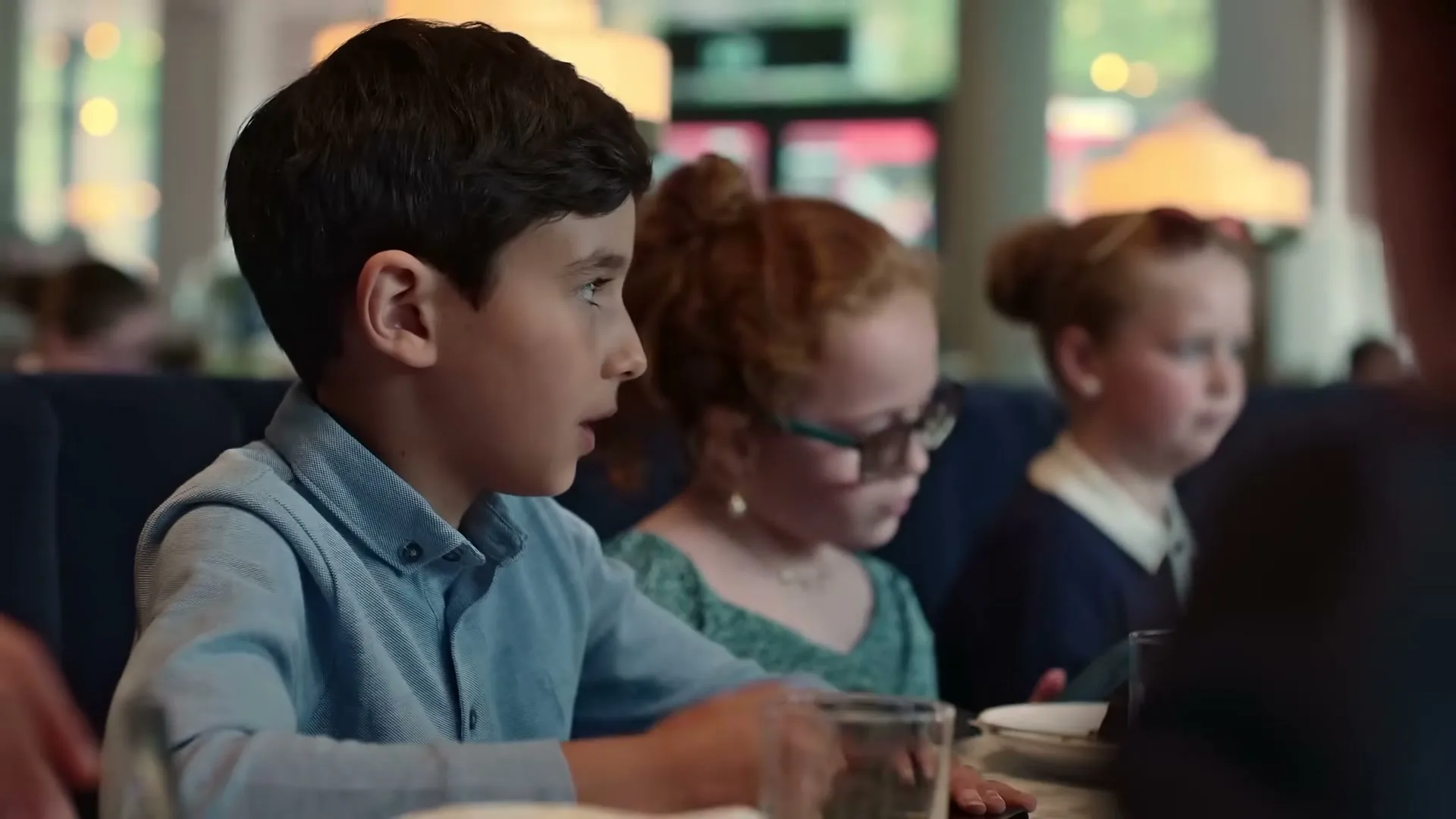 A young boy in a blue shirt sits at a restaurant table with two young girls, looking at something in his hands.