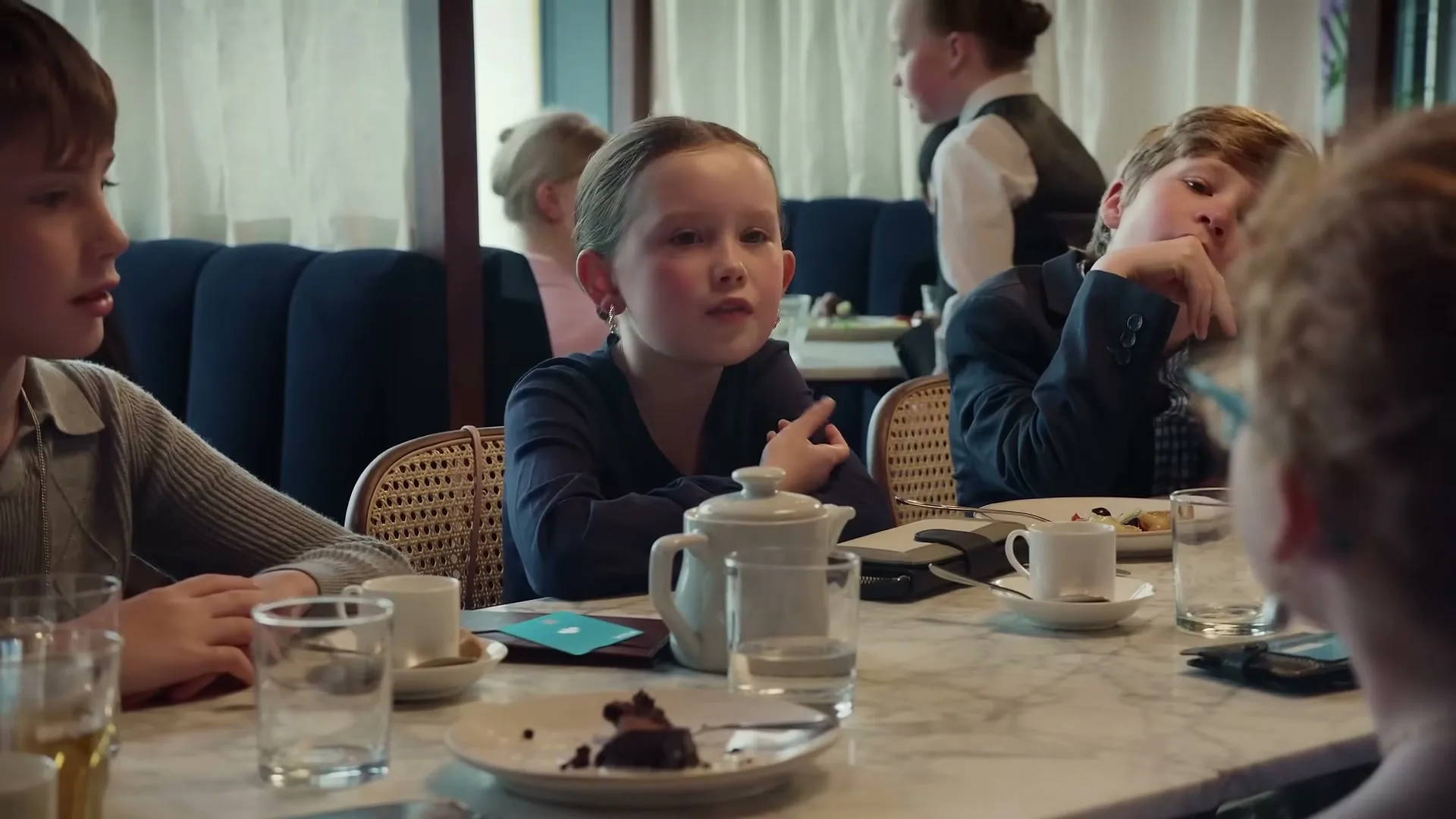 A young girl in a dark top talks at a table with other children in a busy restaurant.