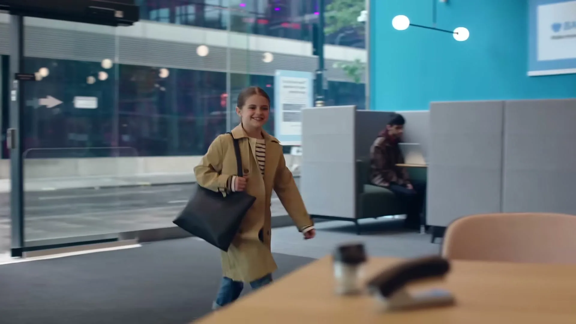 A young girl, dressed in a trench coat and carrying a bag, walks into a modern office building with a smile, as people work in the background.