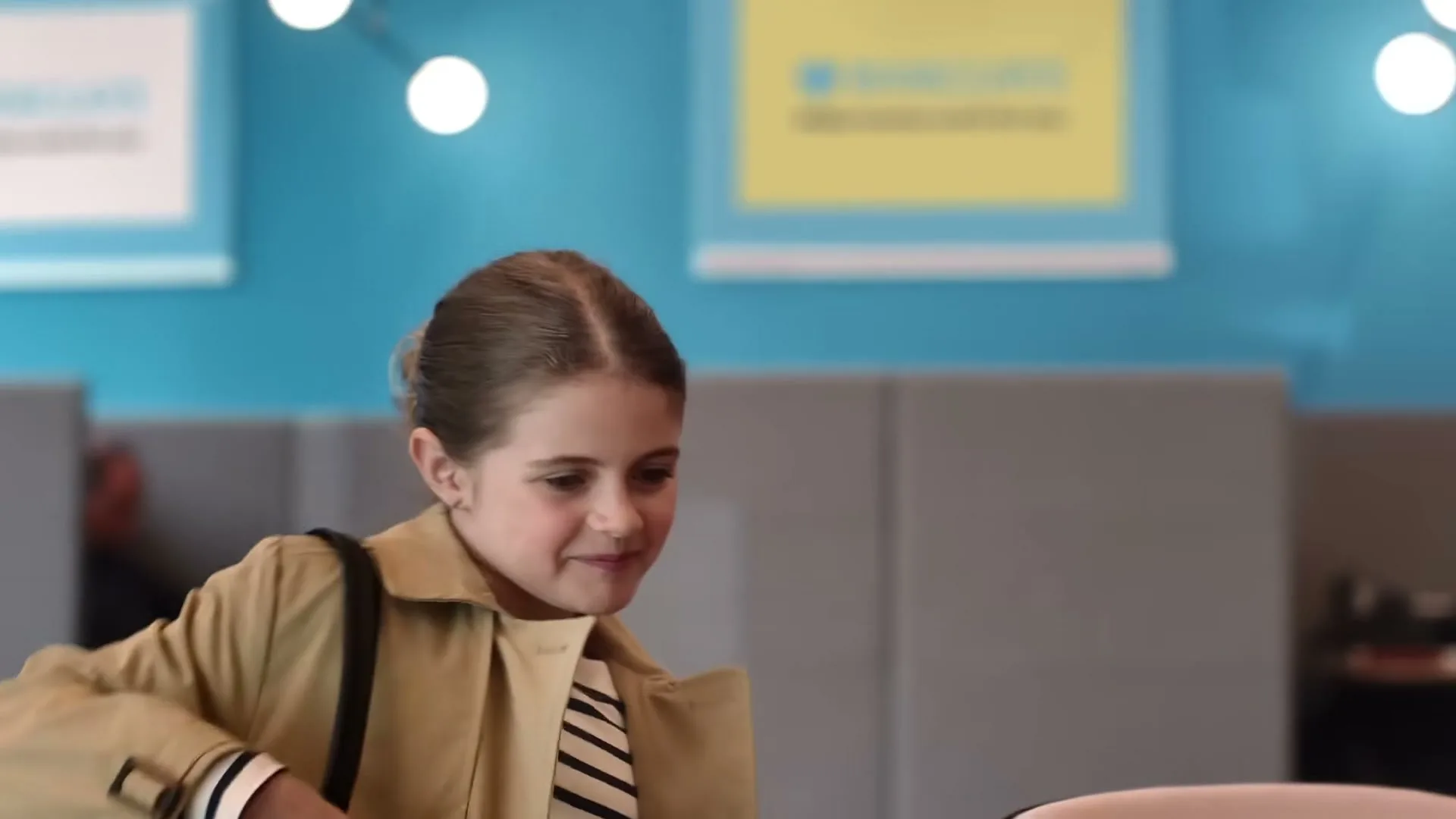 A young girl in a trench coat and striped shirt smiles while holding a bag, looking at someone off-screen in a modern office setting.
