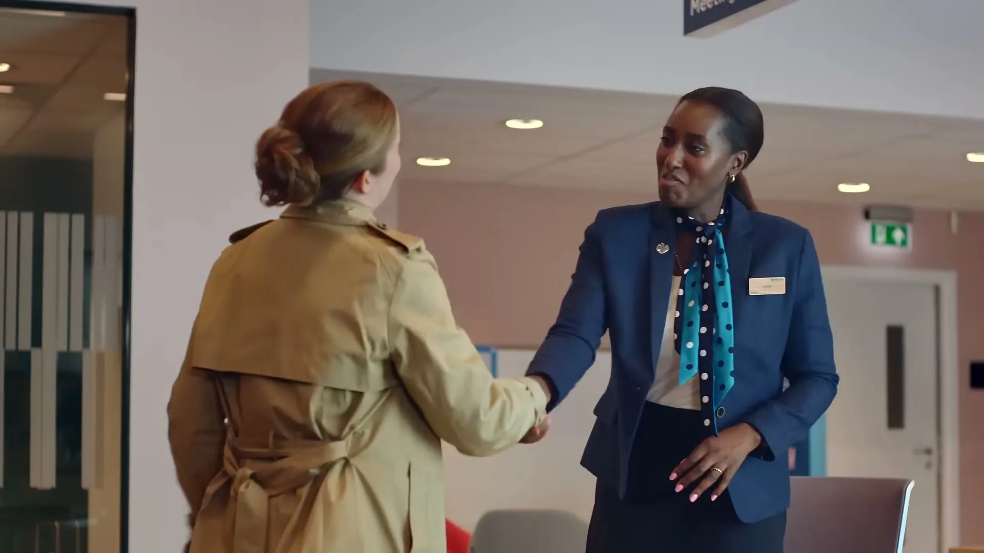 A female bank employee in a blue uniform shakes hands with a female customer in a trench coat in a modern bank lobby.
