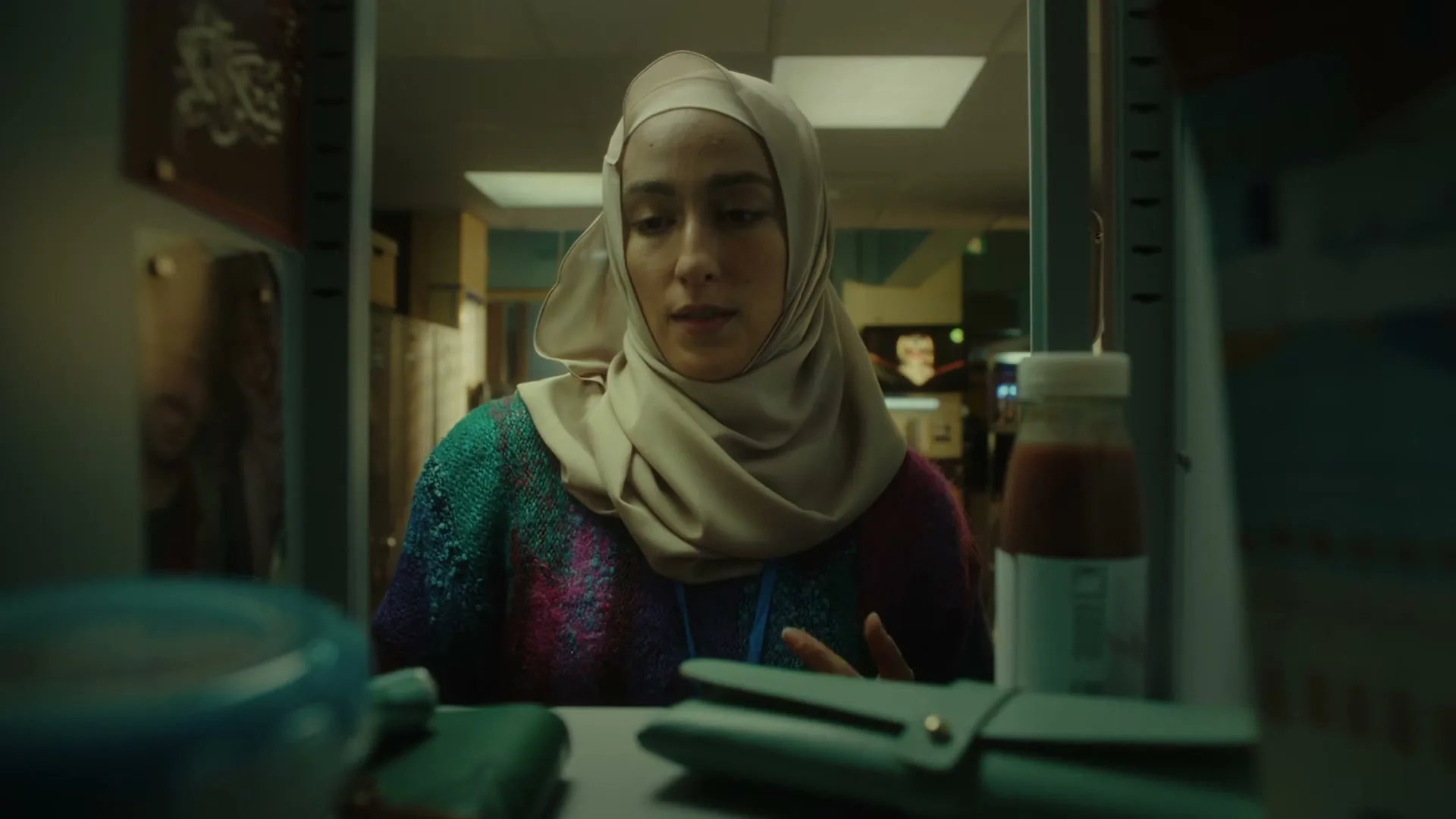 A young woman in a hijab and colorful sweater opens a locker, takes out a red apple, and bites into it, in a school hallway.