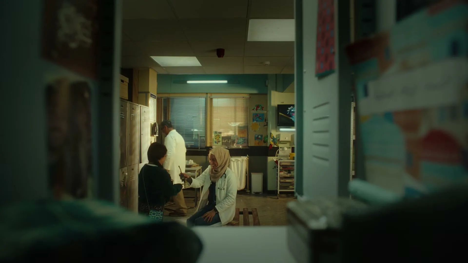 A young boy and a female doctor in a hijab sit on the floor of a medical facility, interacting with each other, as another doctor walks by in the background.