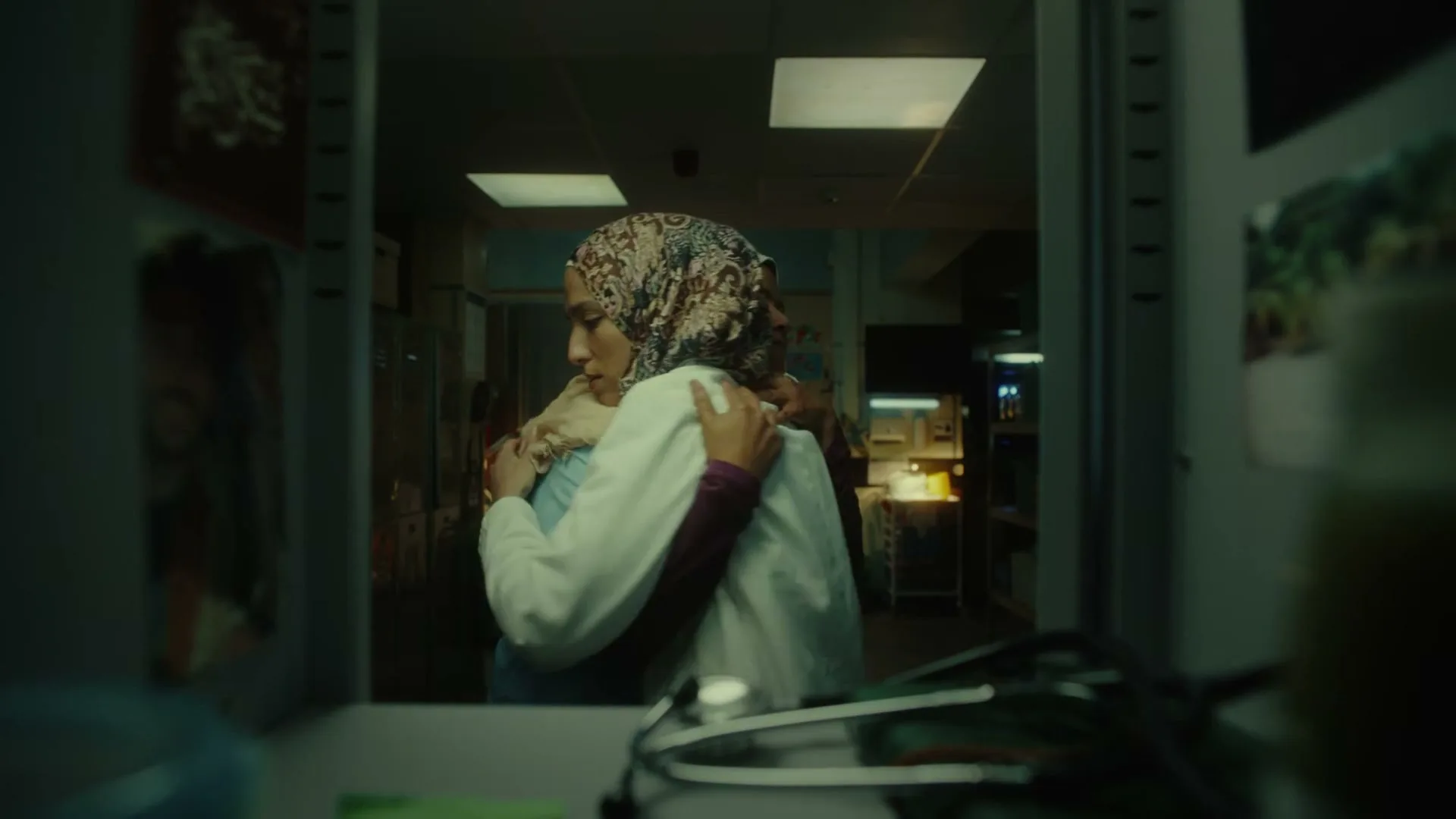 A female doctor in a hijab and white coat opens a locker, then hugs a female colleague in a blue scrub top and hijab, seen through the locker door.