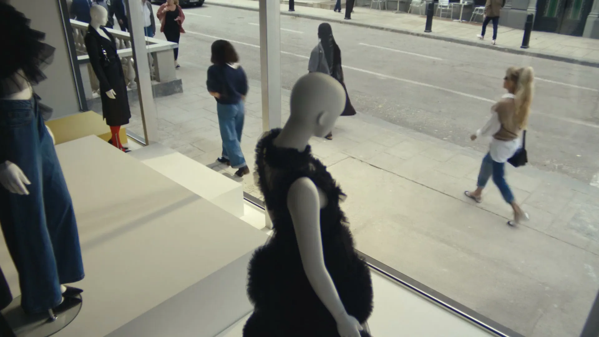A high-angle shot from inside a store window shows a mannequin in a black dress, with people walking past on the street outside.