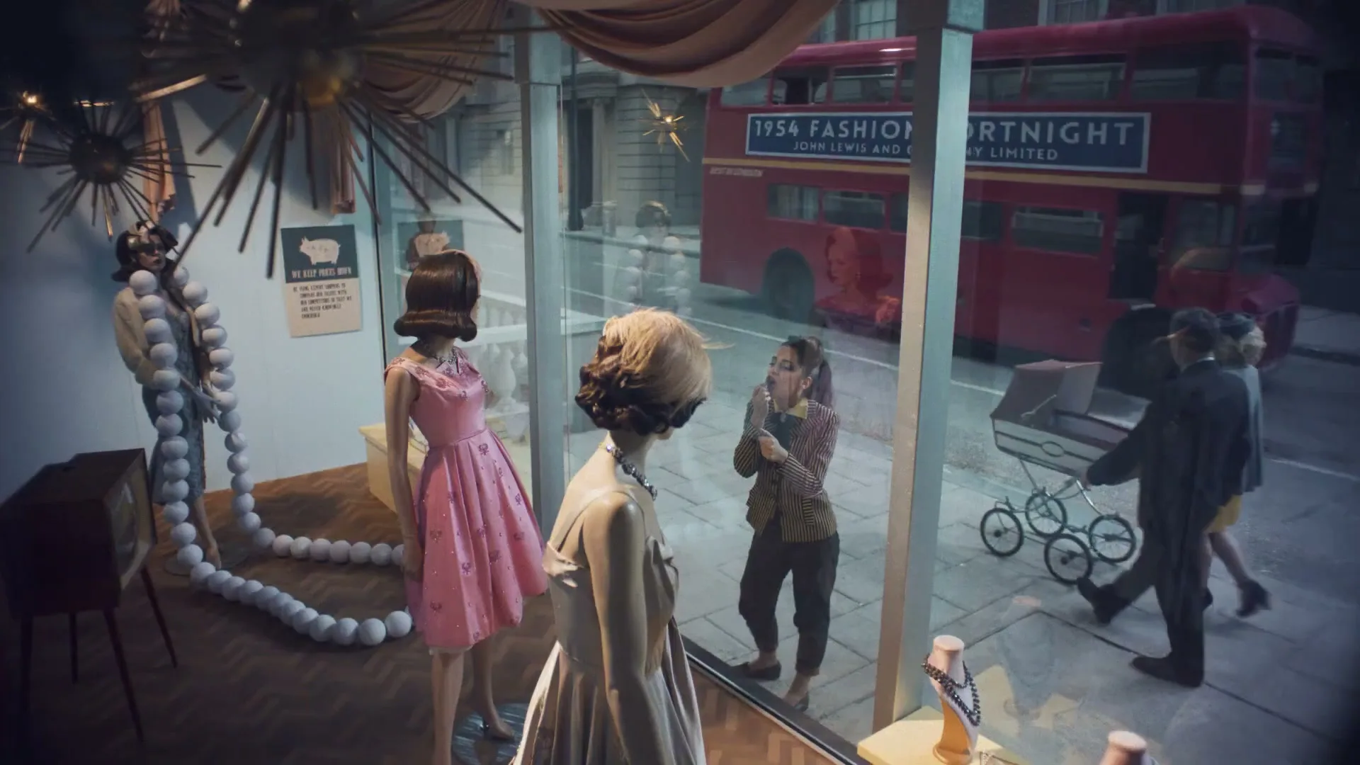 A young woman in a striped jacket and dark pants looks into a shop window, admiring the dresses on mannequins, while a red double-decker bus passes by on the street.