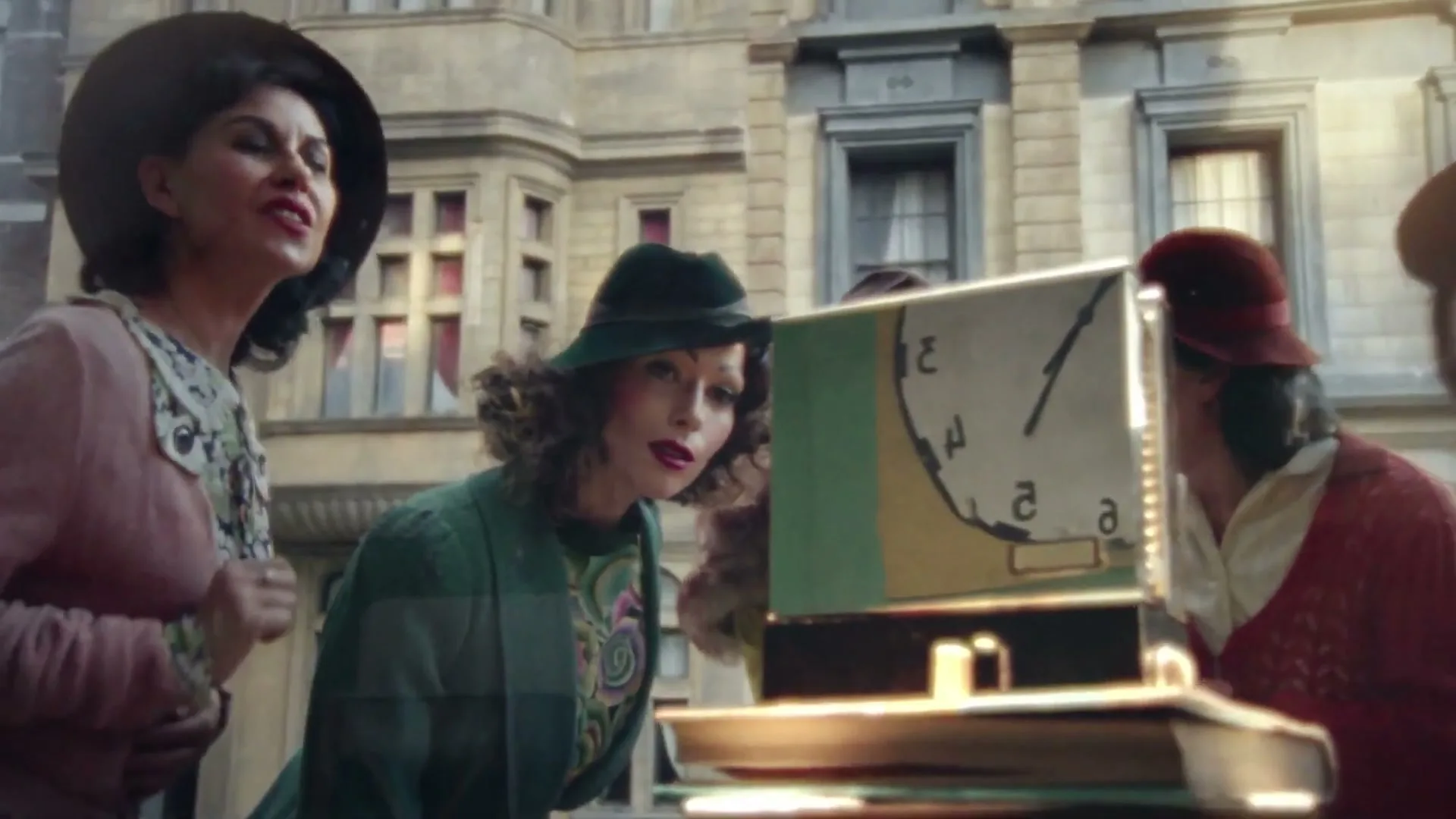 Four women in vintage clothing look intently at a clock display in a shop window, with a building facade in the background.