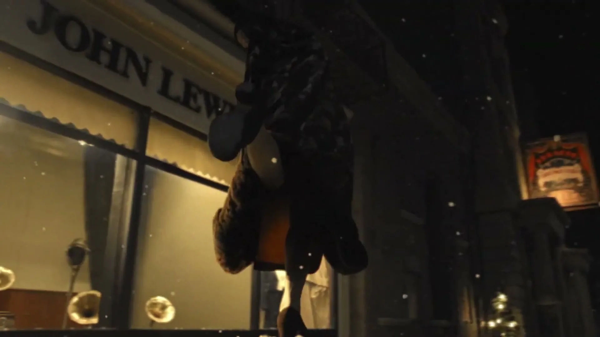 A person in a brown coat and white pants is seen from a low angle, walking past a John Lewis & Co. store at night, with snow falling.