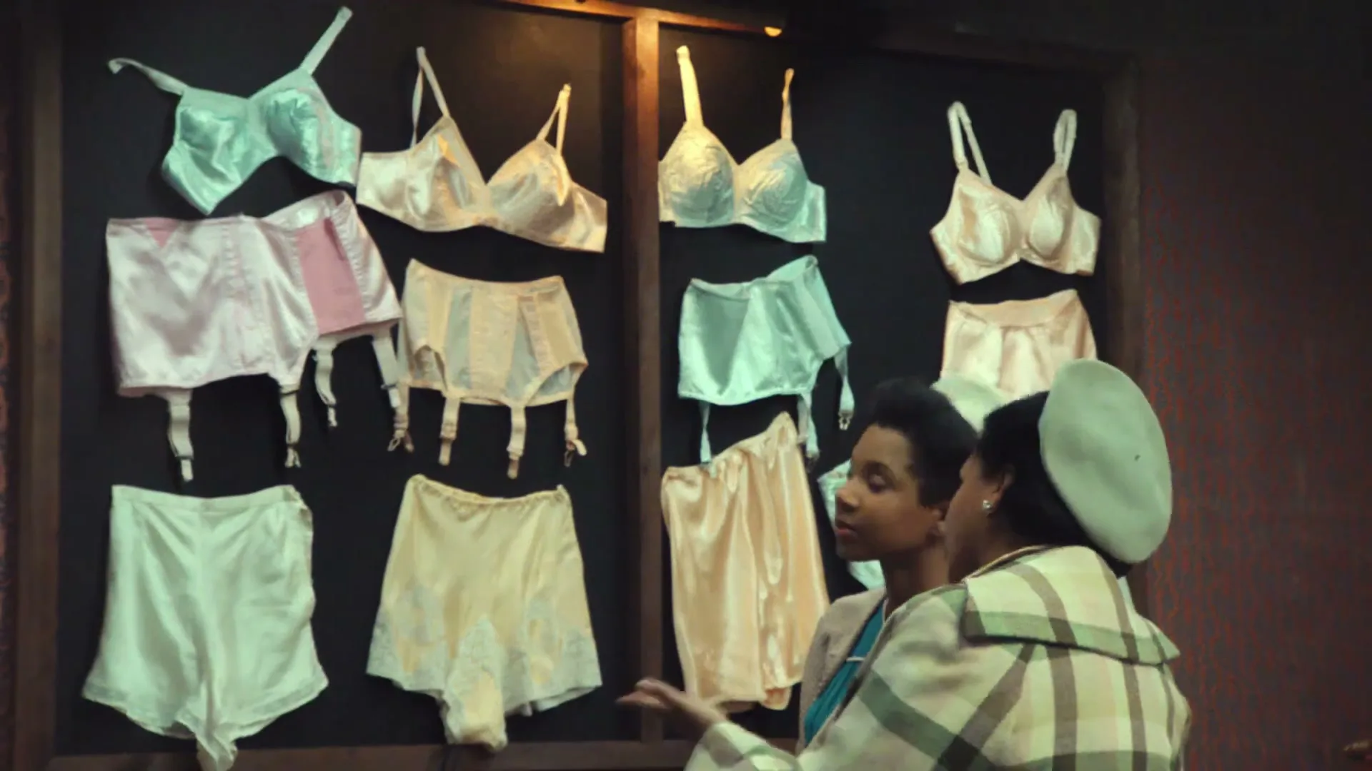 Two women, one in a plaid coat and beret, the other in a blue dress, look at lingerie displayed on a dark wall in a department store.