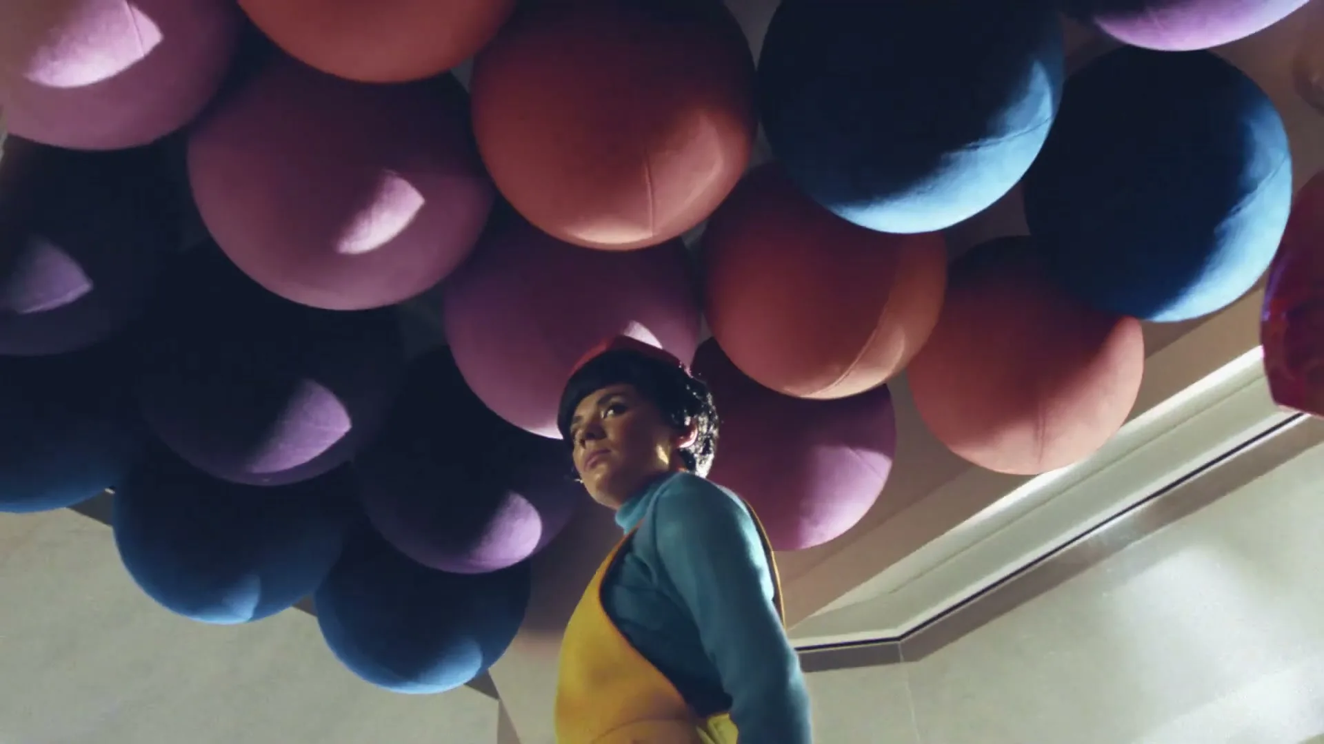 A person with short dark hair and a red hat looks up, surrounded by large colorful spheres hanging from the ceiling in an interior space.