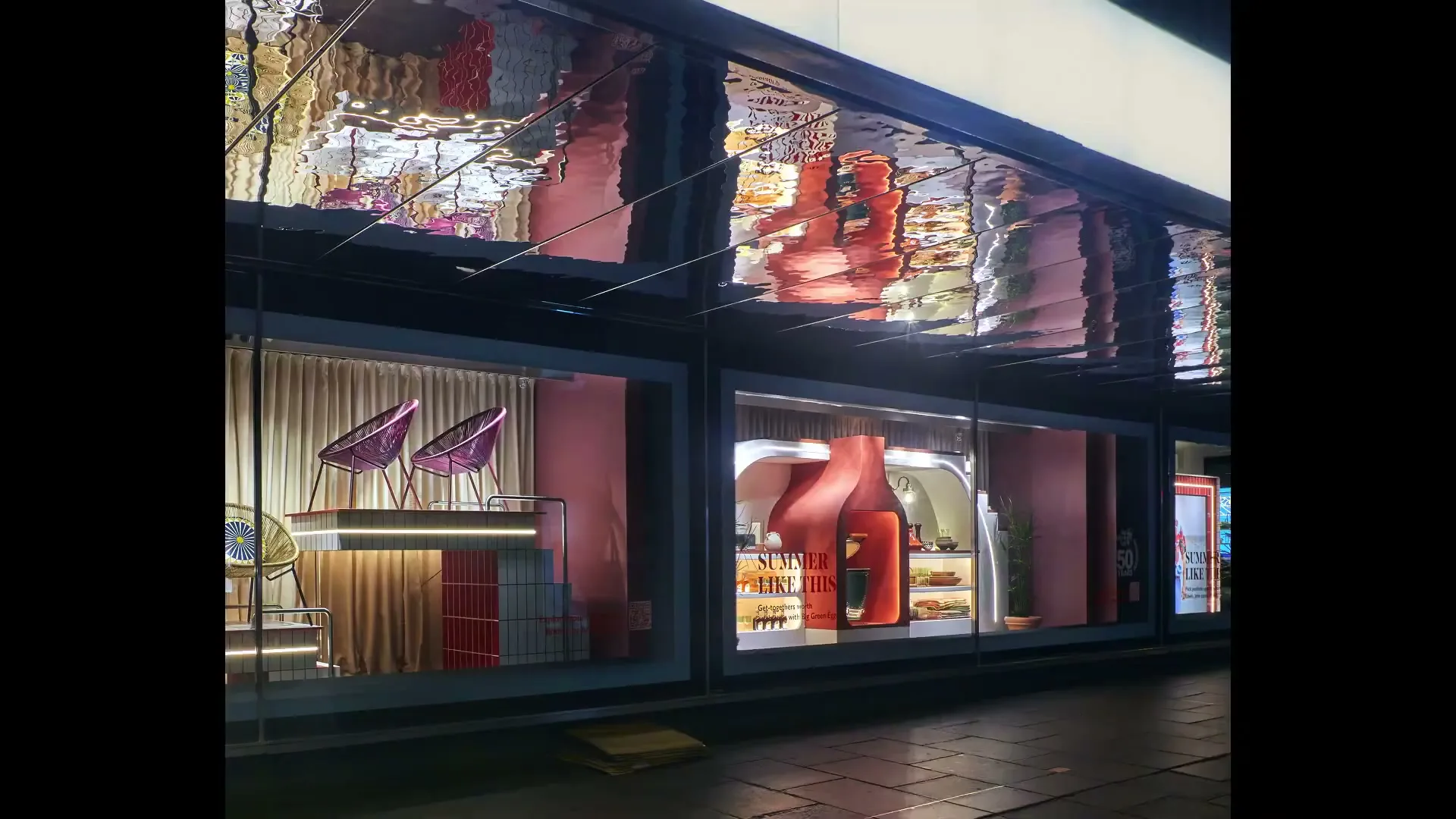 A wide shot of a department store window display at night, featuring various home goods and a large bottle-shaped cooler.
