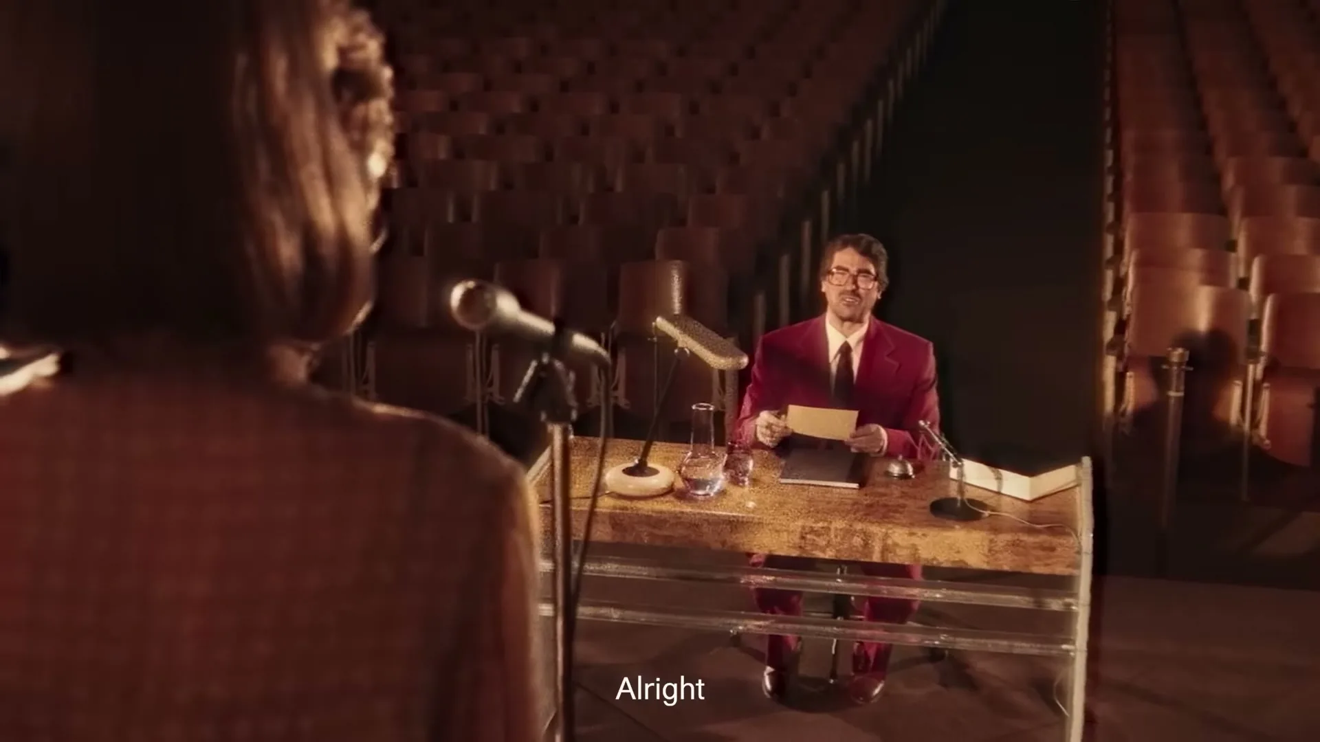 A woman with brown hair stands at a microphone, looking at a man in a red suit and glasses who sits at a desk in an empty auditorium.