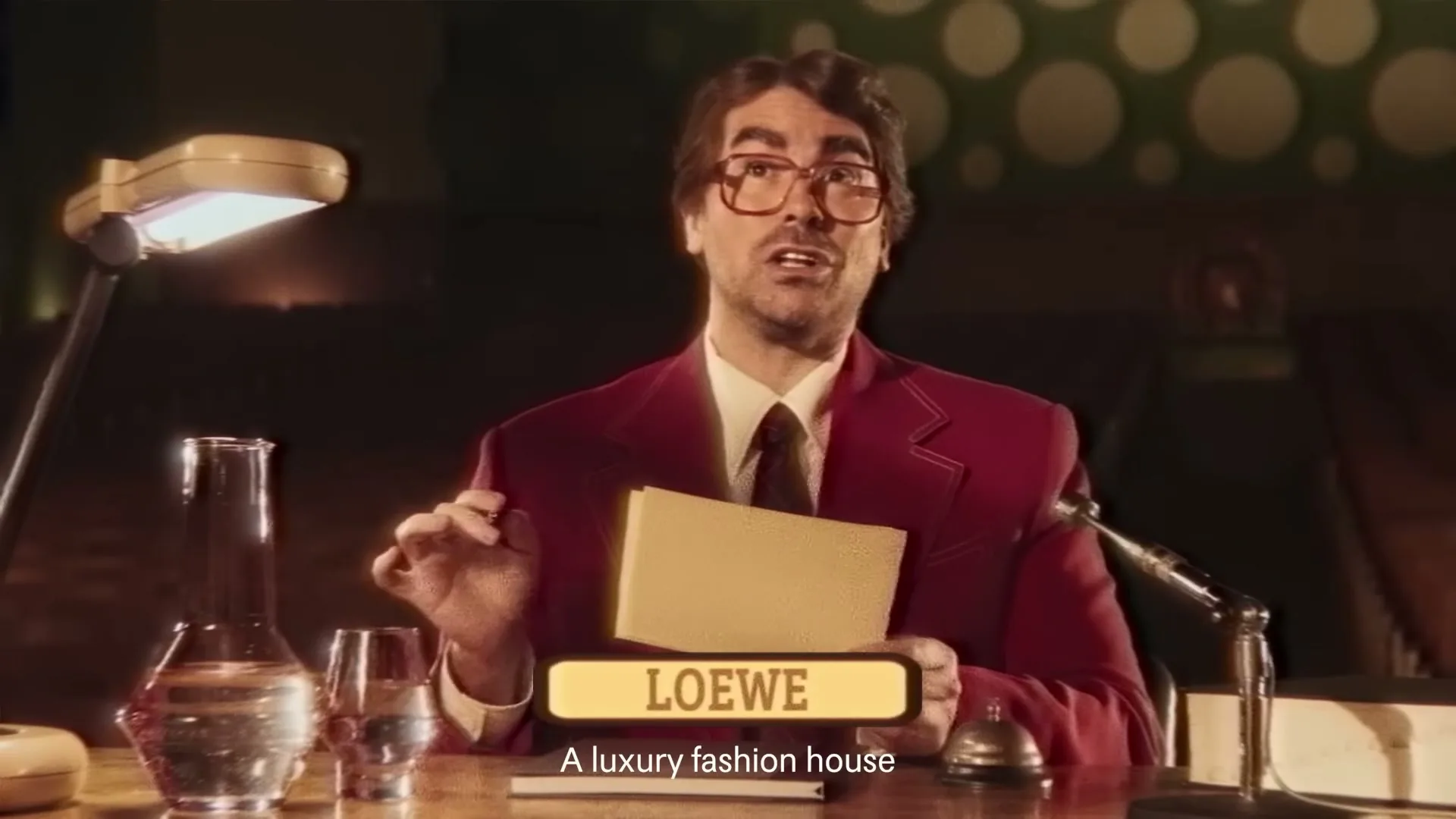 A man in a red suit and glasses sits at a desk, holding papers and speaking, with a "LOEWE" graphic on screen.