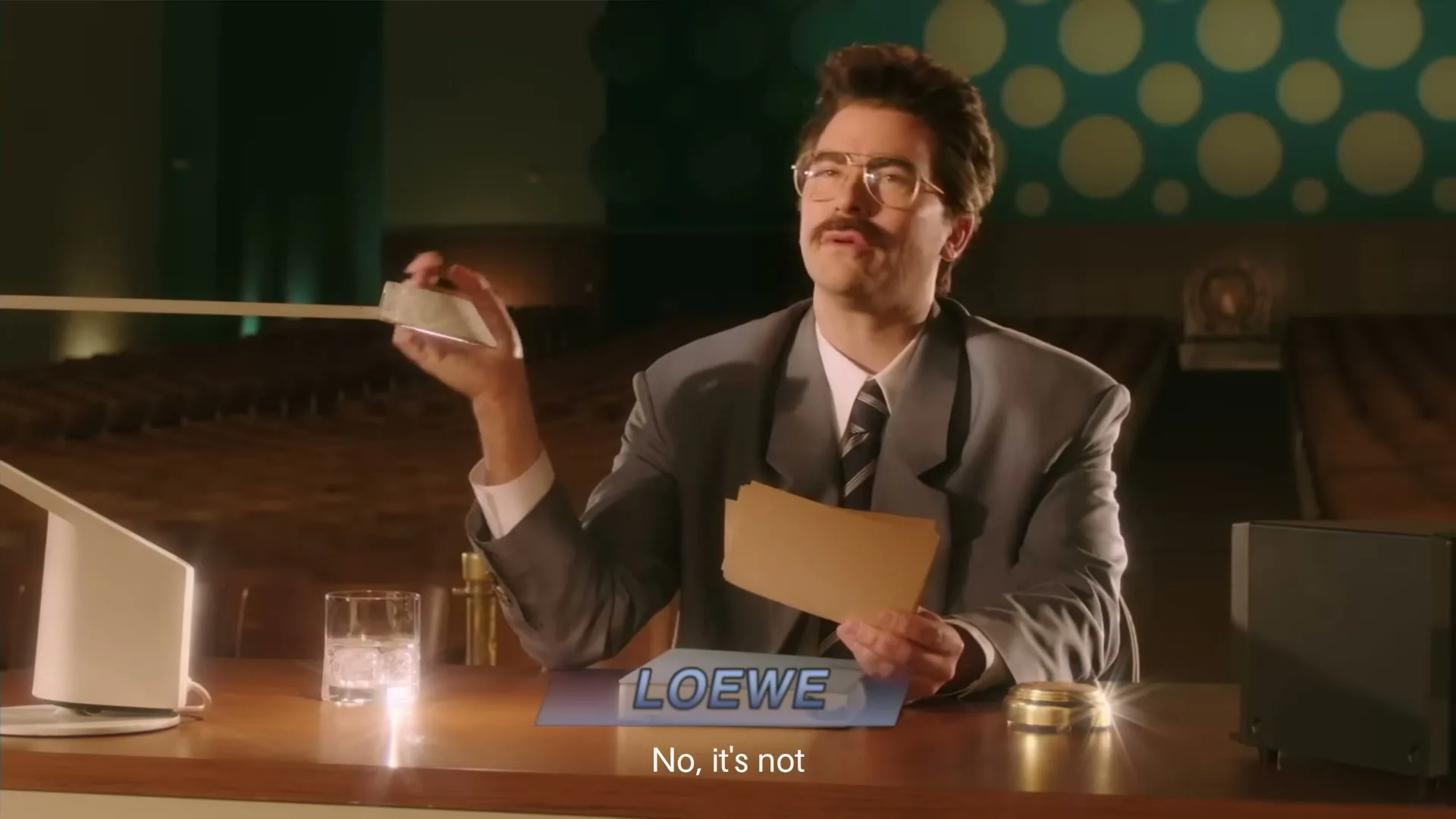 A man with a mustache and glasses, wearing a suit, sits at a desk with a "LOEWE" sign, holding papers and a lamp.