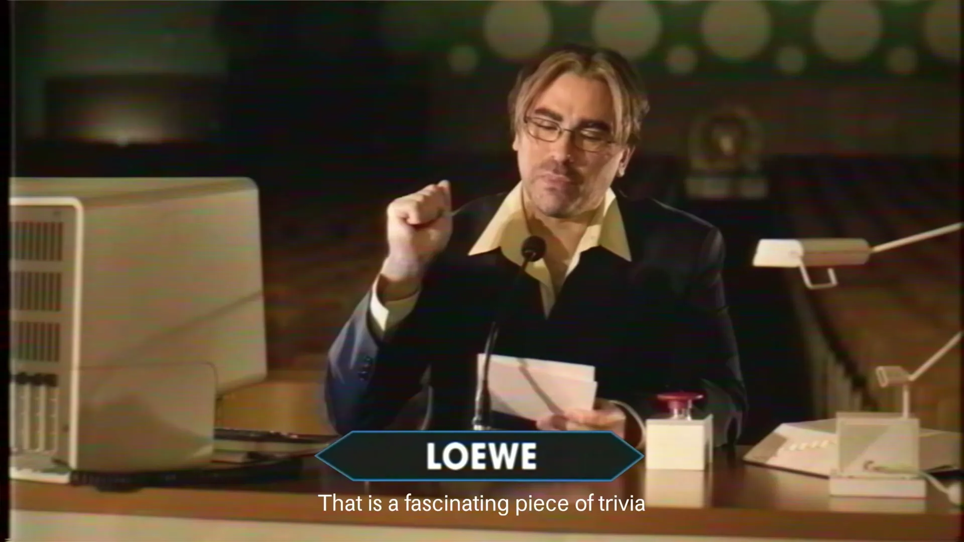 A man with glasses and a suit sits at a desk, holding papers and gesturing, with a computer and lamp beside him in a dimly lit room.