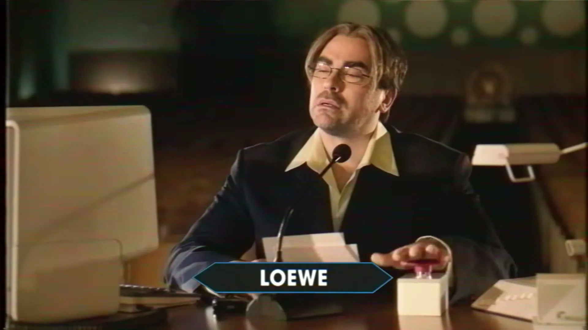 A man with glasses and a suit sits at a desk with a microphone, looking confused, in a dimly lit room.