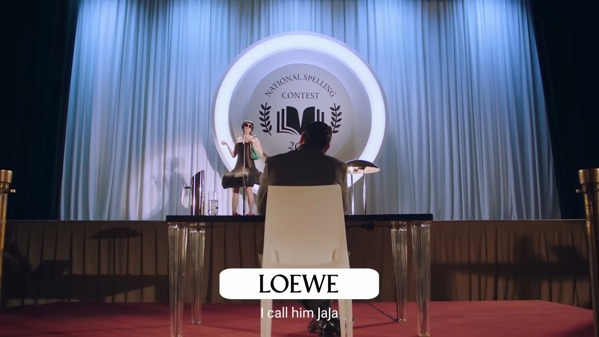 A woman in a green dress and sunglasses stands at a microphone on a stage, facing away from the camera, while a man in a suit sits at a clear table with his back to the camera, at a spelling bee.