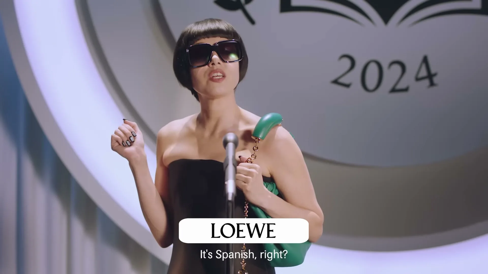 A woman with a bob haircut and sunglasses stands at a microphone, holding a green handbag, with "2024" visible in the background.