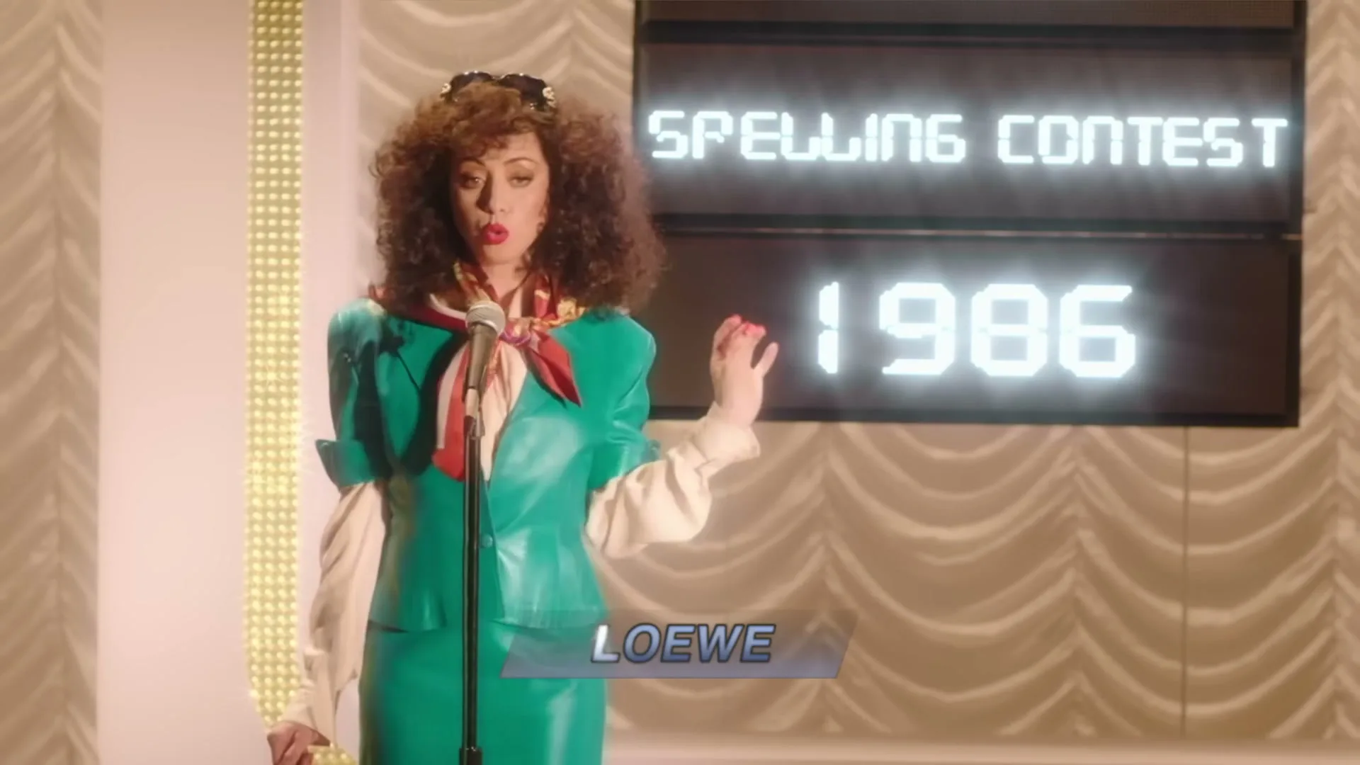 A woman with big curly hair, wearing a green leather suit and a scarf, stands at a microphone on a stage with a "Spelling Contest 1986" sign.