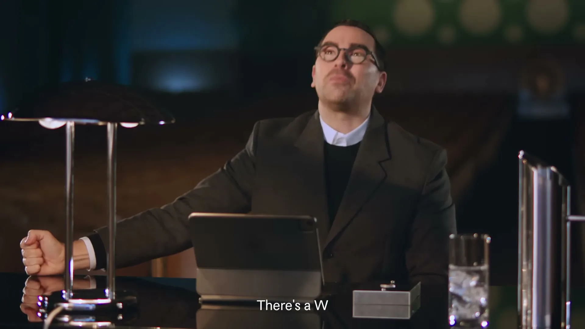 A man in a suit and glasses sits at a desk with a lamp and tablet, looking up and then smiling, in an interior setting.