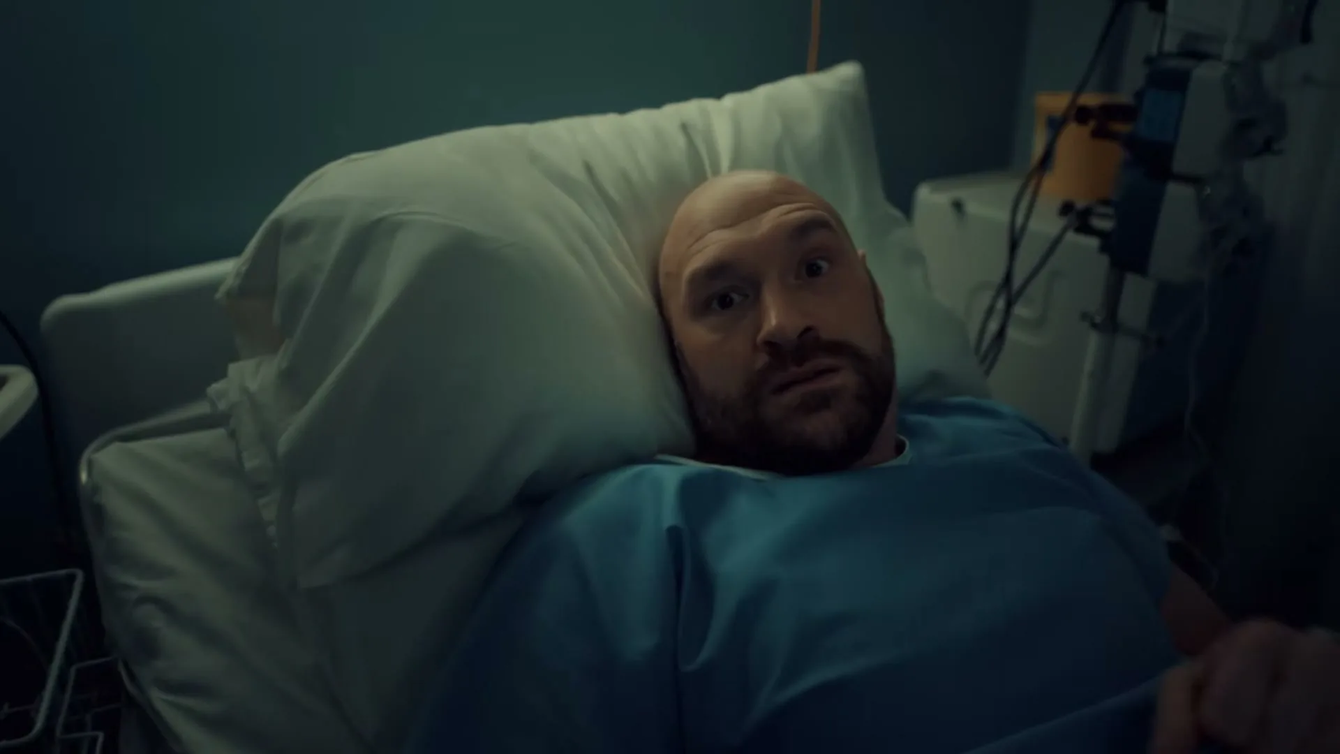 A bald, bearded man in his 30s lies in a hospital bed, looking surprised, in a dimly lit hospital room.