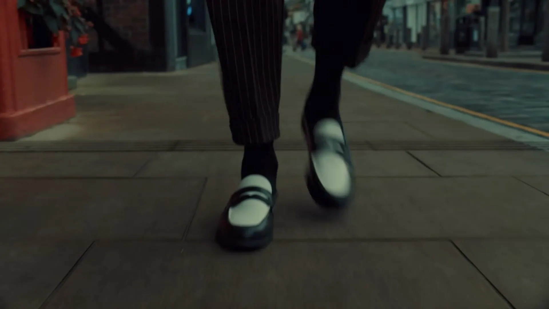 A person's feet in black and white loafers and pinstripe pants walk along a paved sidewalk next to a street.