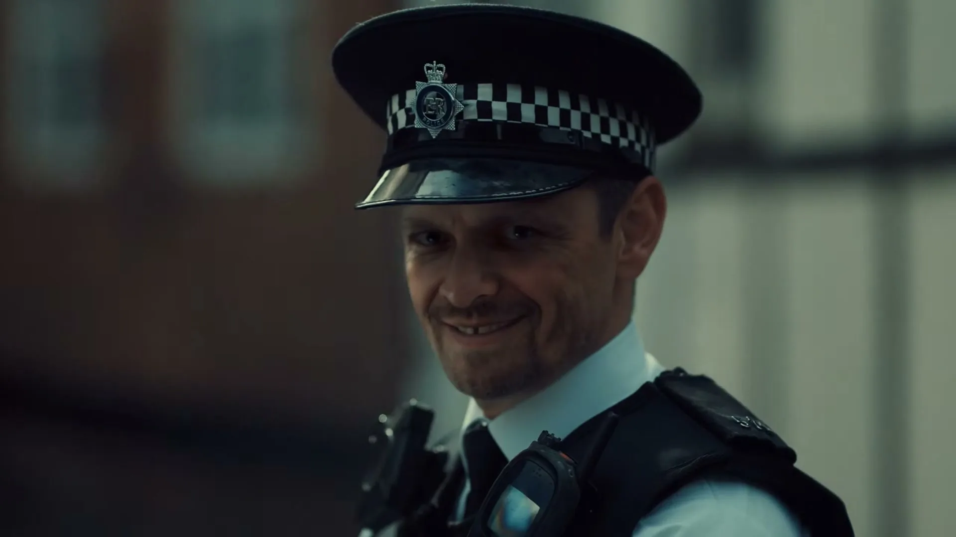 A male police officer in uniform smiles subtly, looking directly at the camera in an urban setting.