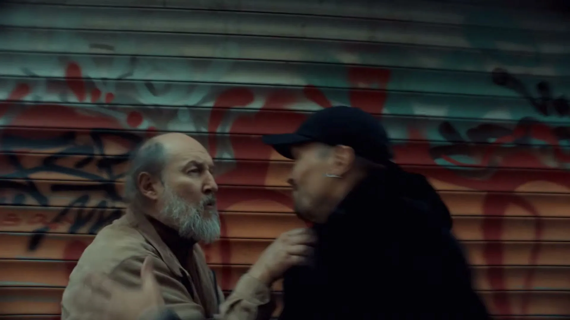 Two men, one older with a white beard and a younger man in a black cap, are in a physical altercation against a graffiti-covered metal wall.