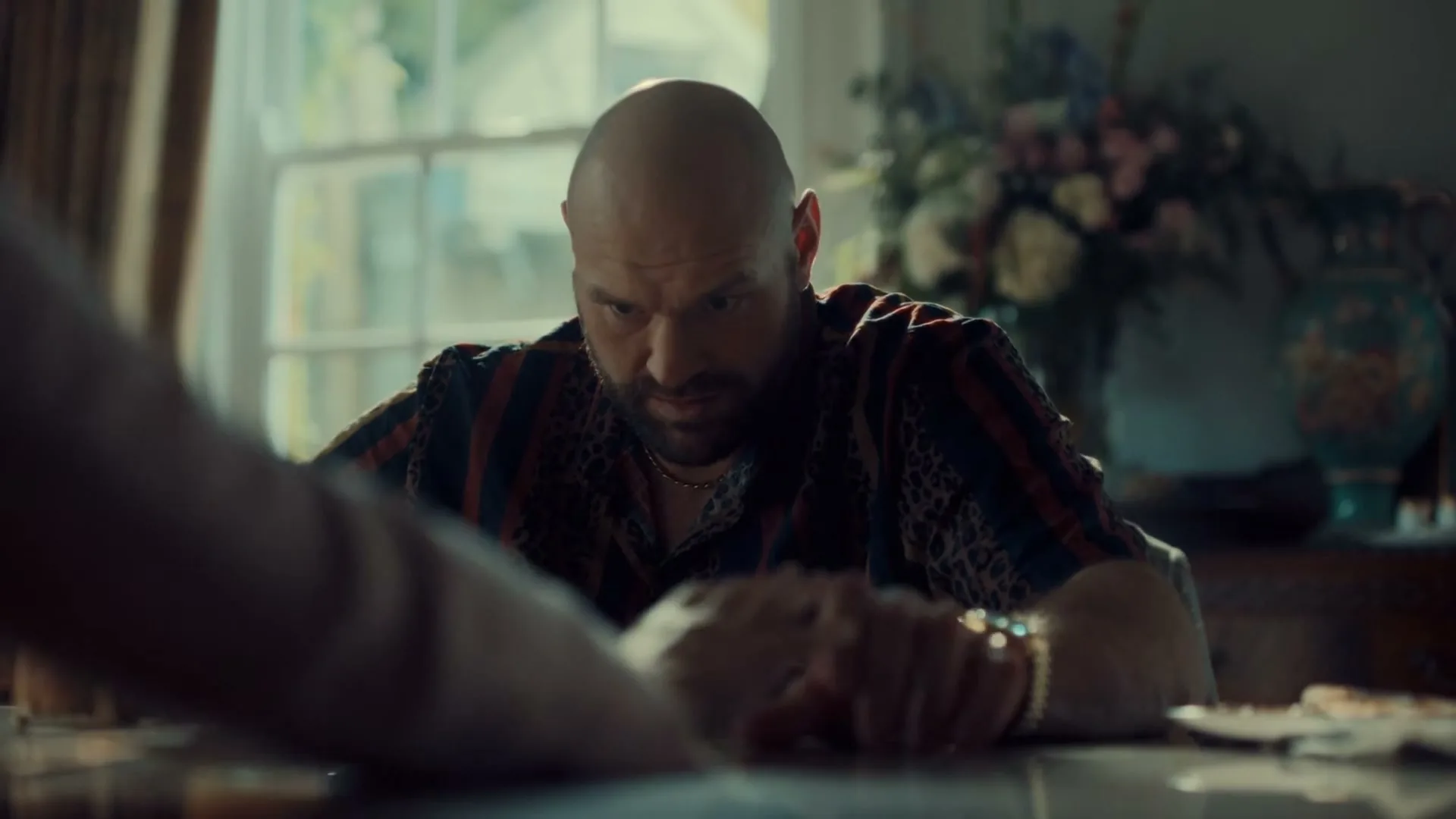 A bald man with a beard, wearing a patterned shirt and gold jewelry, sits at a dining table, looking down intently, then glances up.