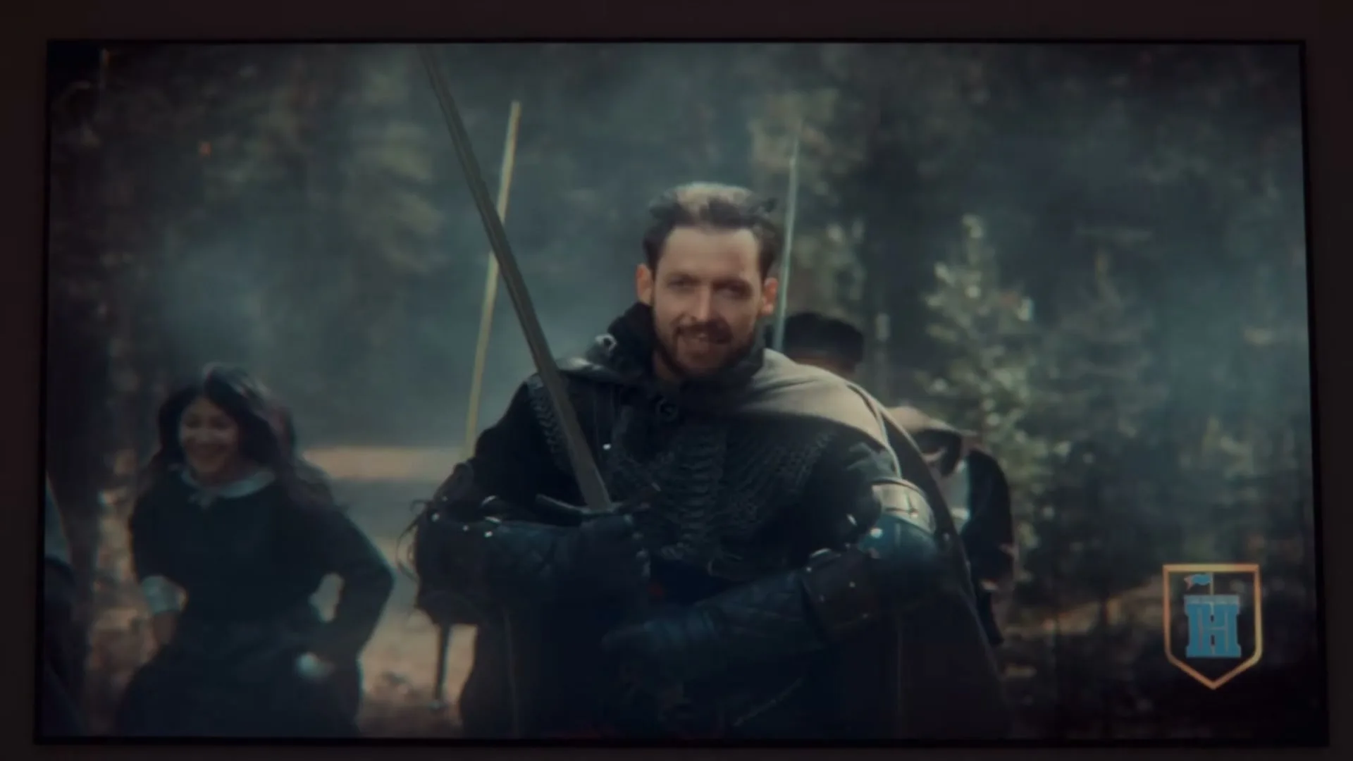 A man in medieval armor with a sword smiles, then the scene transitions to an animated man tied up, on a screen in an interior setting.