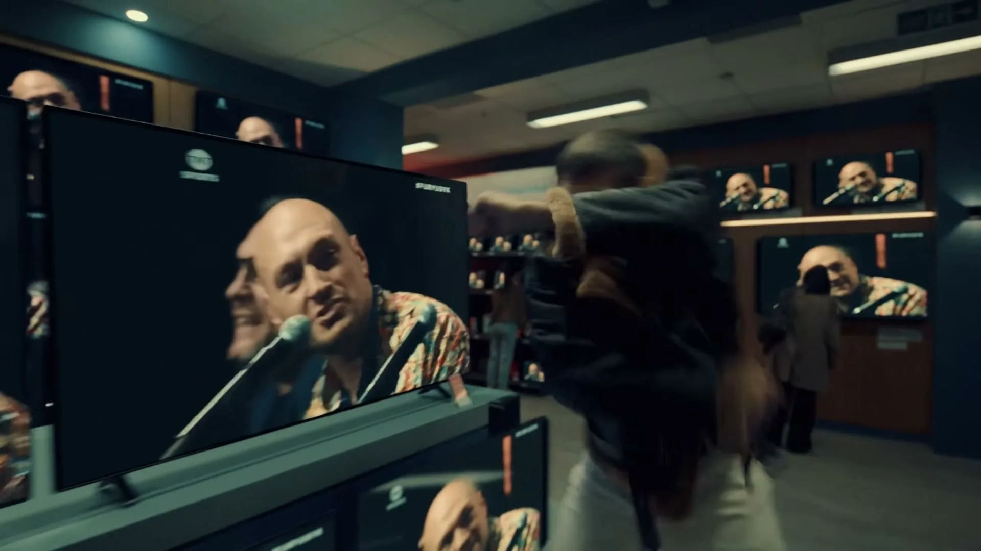 A man in a bomber jacket runs through an electronics store, dodging multiple TVs displaying a bald man singing into a microphone.