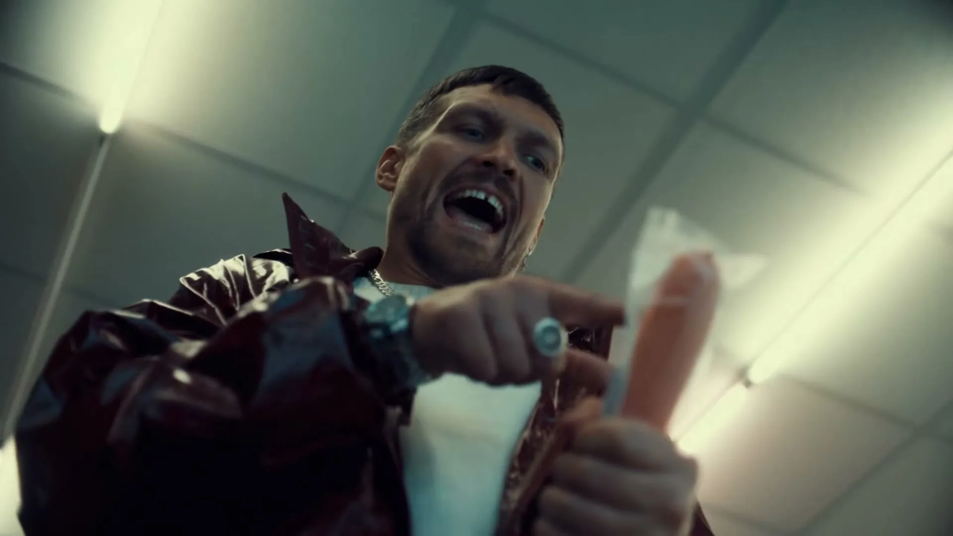 A man in a red jacket and white shirt holds a sausage in a plastic bag, pointing at it with an angry expression in a brightly lit room.