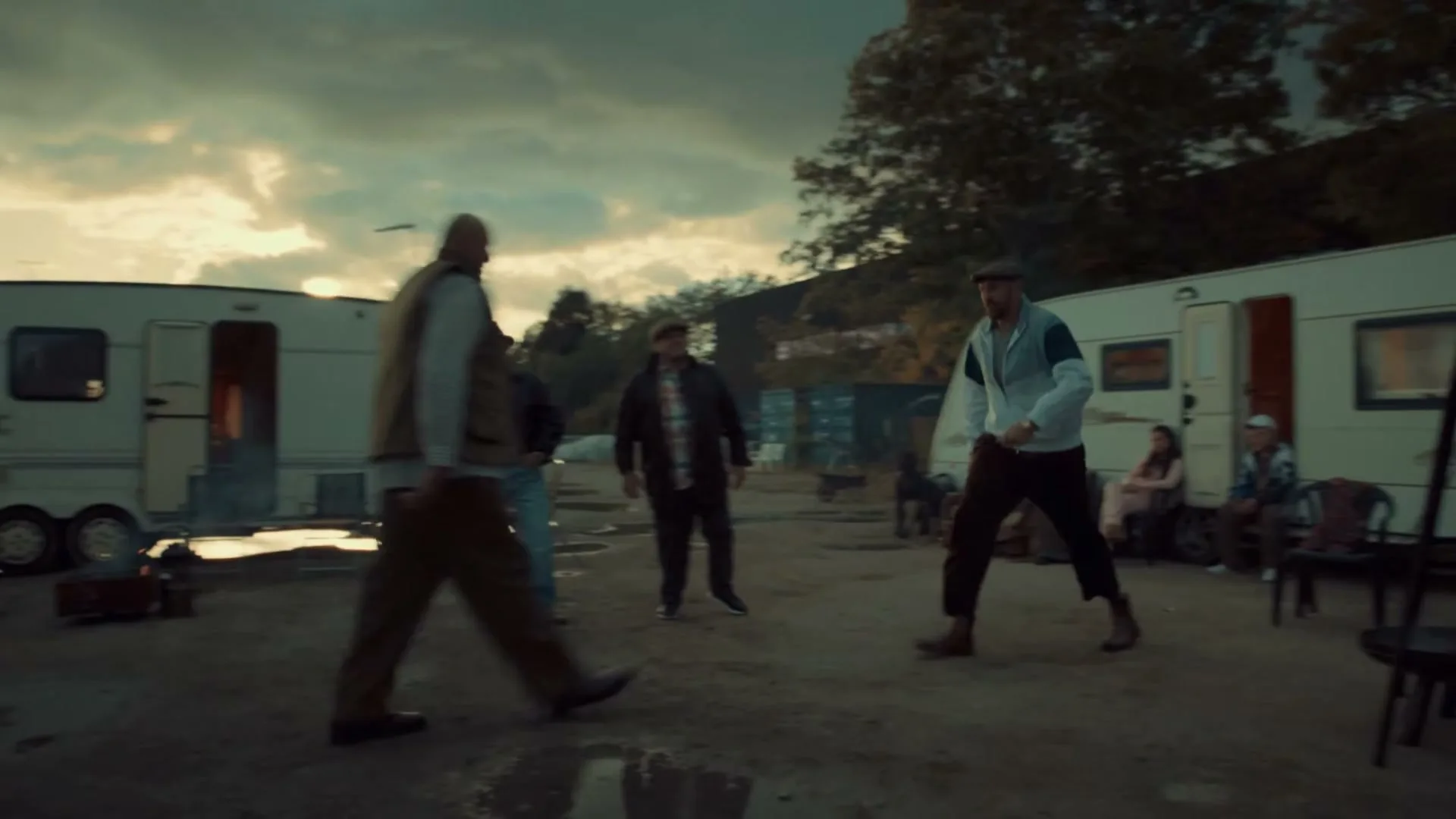 A group of men in casual attire gather in a trailer park under a dramatic sky, with one man walking past the camera.