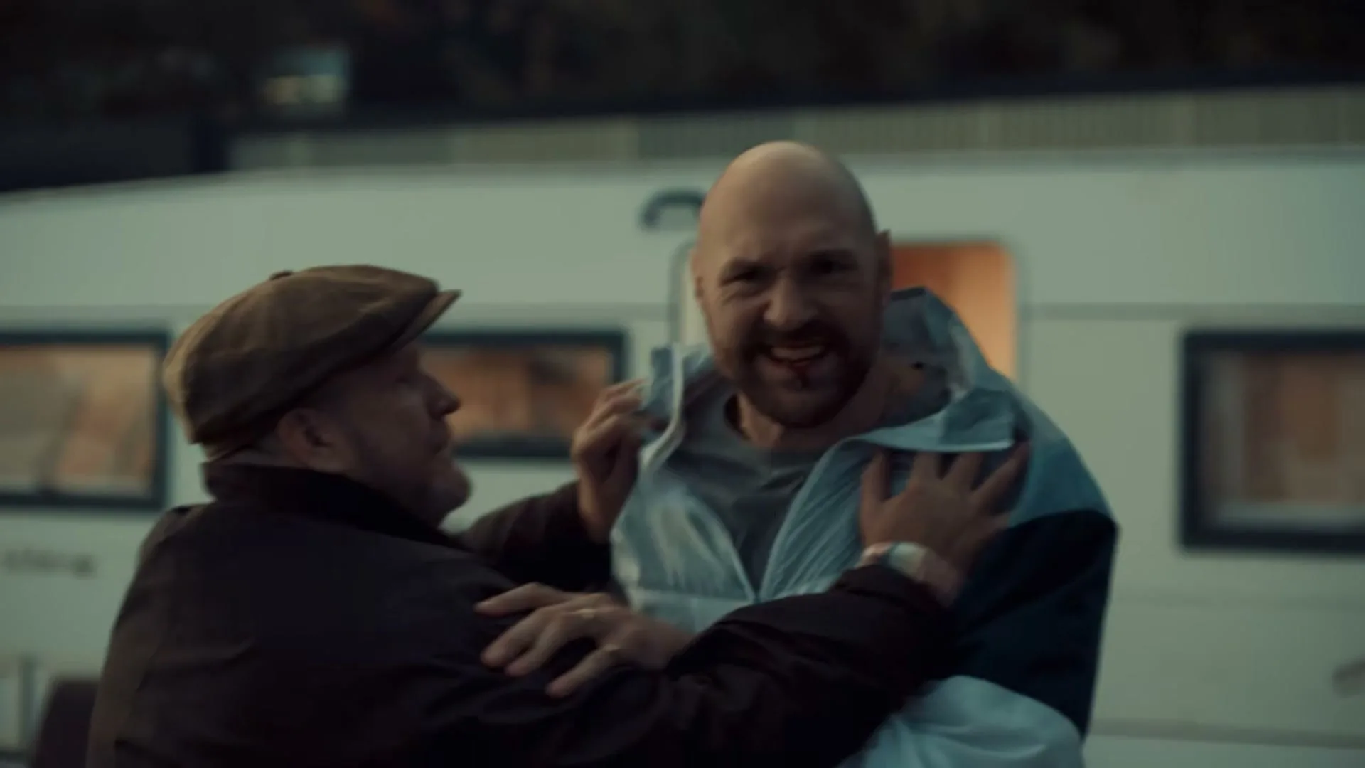 A bald man in a light blue and white jacket is held by an older man in a brown cap and jacket, both looking distressed outside a caravan.
