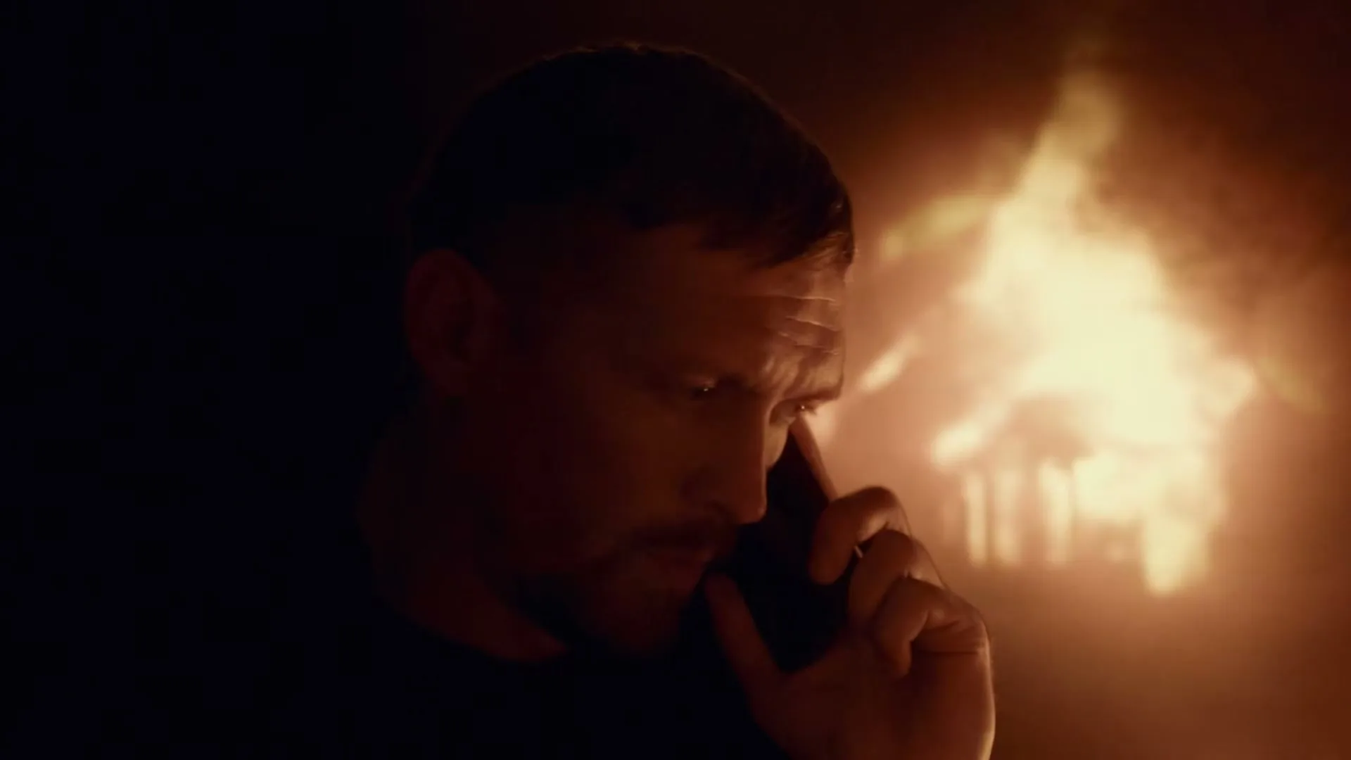 A man with a beard holds a phone to his ear, his face partially illuminated by a warm glow from the right, in a dark setting.