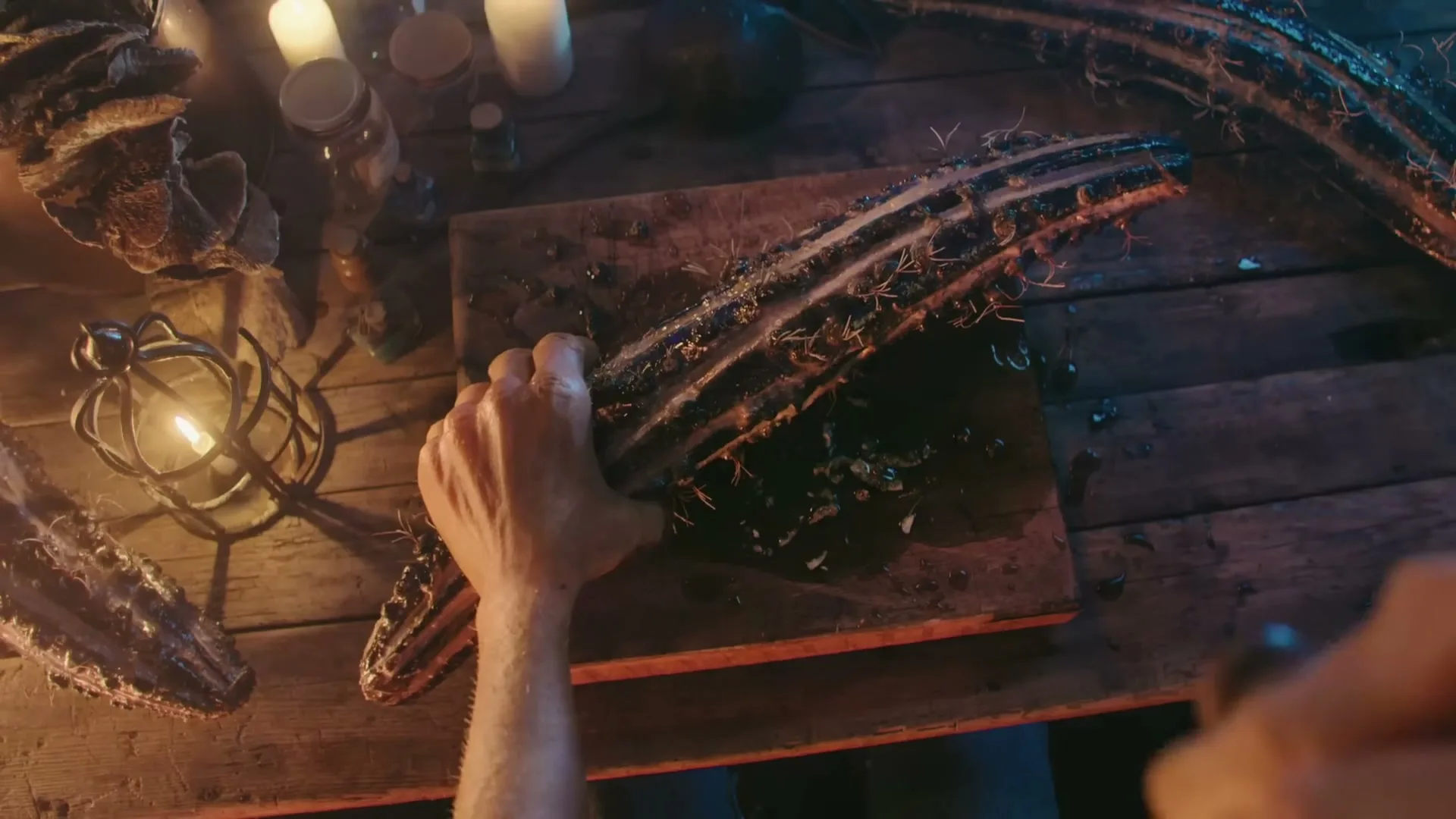 A man's hands are seen from an overhead perspective, cutting a strange, spiky vegetable on a wooden cutting board in a dimly lit, rustic setting.