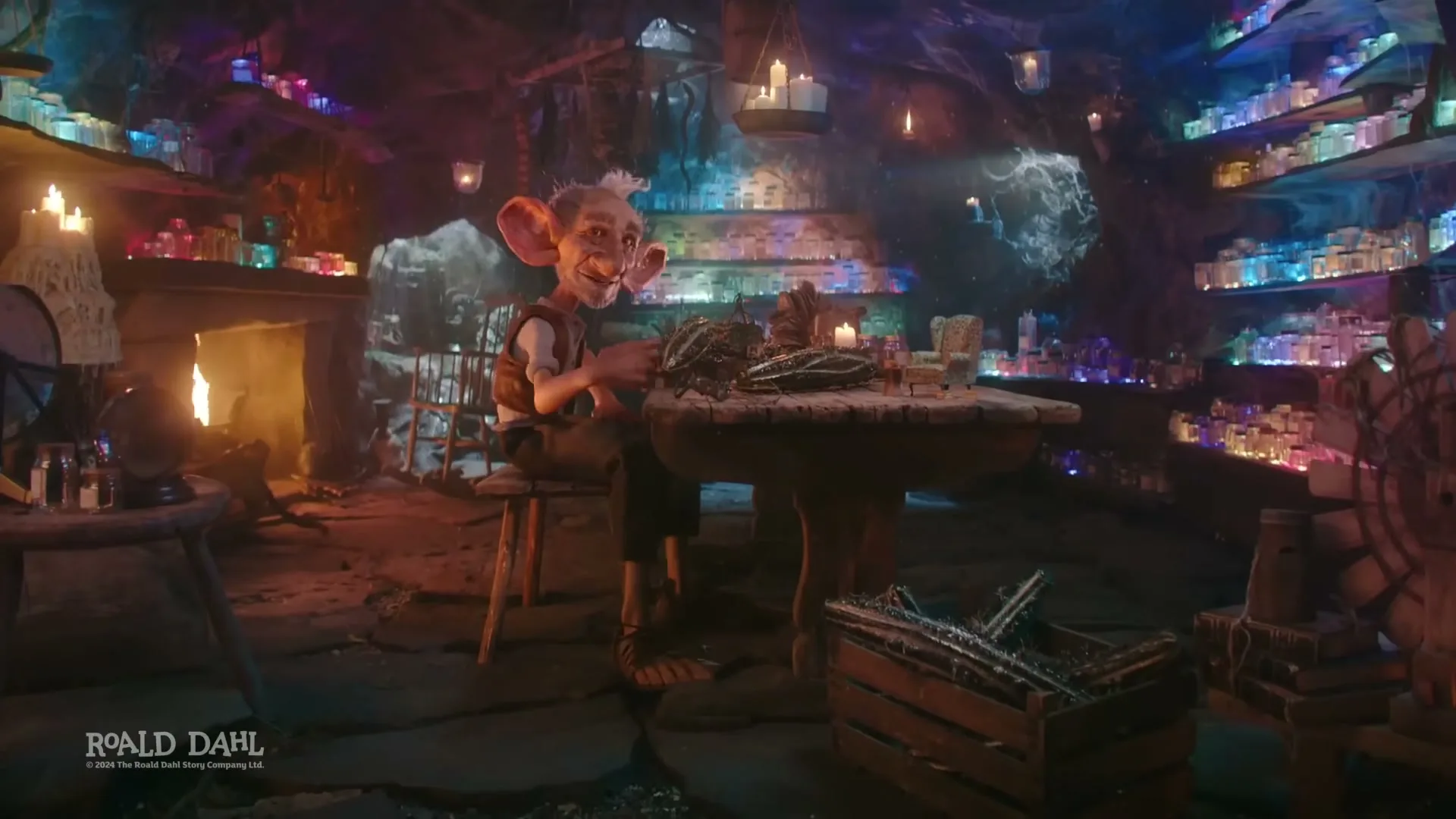 A small, old man with large ears and white hair sits at a wooden table, surrounded by shelves filled with colorful jars in a dimly lit, magical cave.