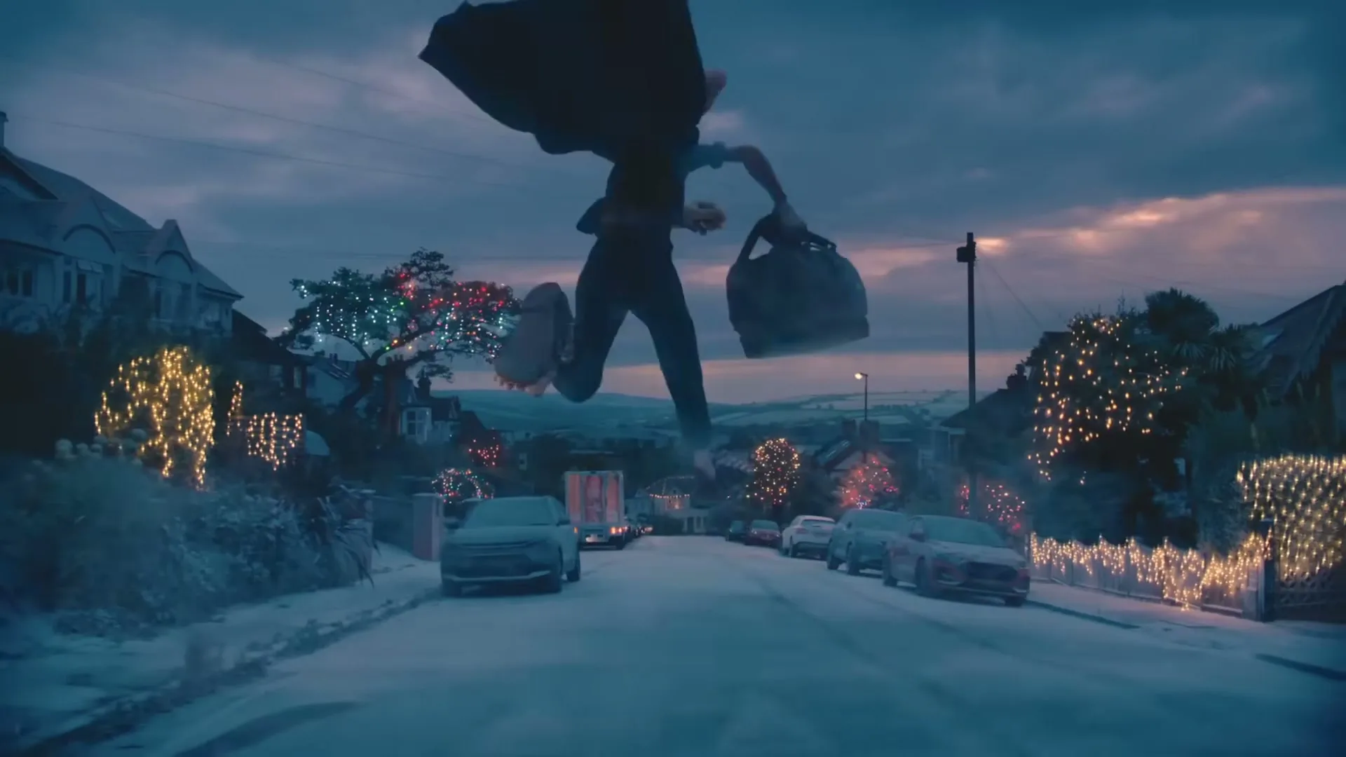 A giant person runs down a snow-covered street lined with houses decorated with Christmas lights, carrying a large bag.