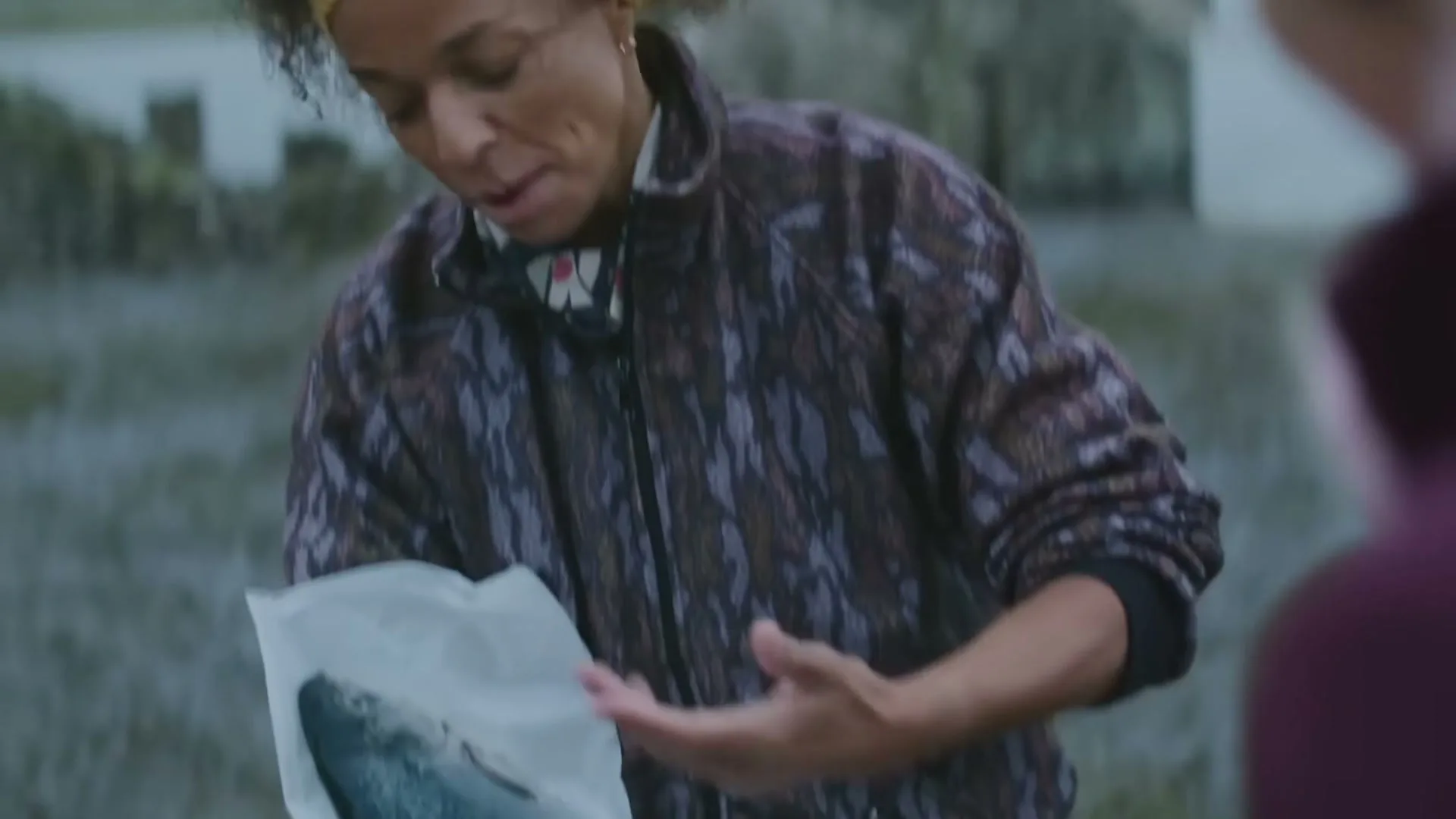 A woman with curly hair and a yellow headband smiles while holding a fresh fish wrapped in paper outdoors, with a stone building in the background.