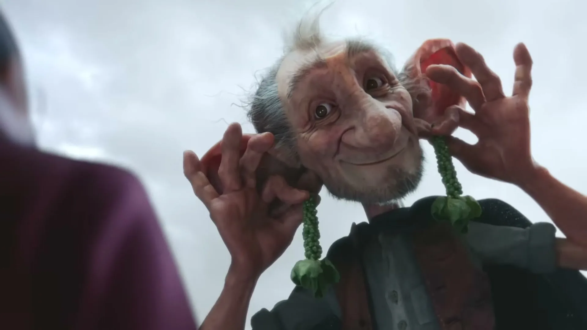 A giant-like creature with large ears and a friendly smile looks down at someone, holding green plant-like earrings to its ears.